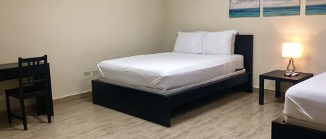 A bedroom with a bed, a desk, a chair, and a nightstand with a lamp.