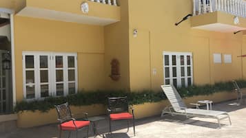 Un edificio amarillo con ventanas de marco blanco, un balcón con barandilla blanca y asientos exteriores con cojines rojos.