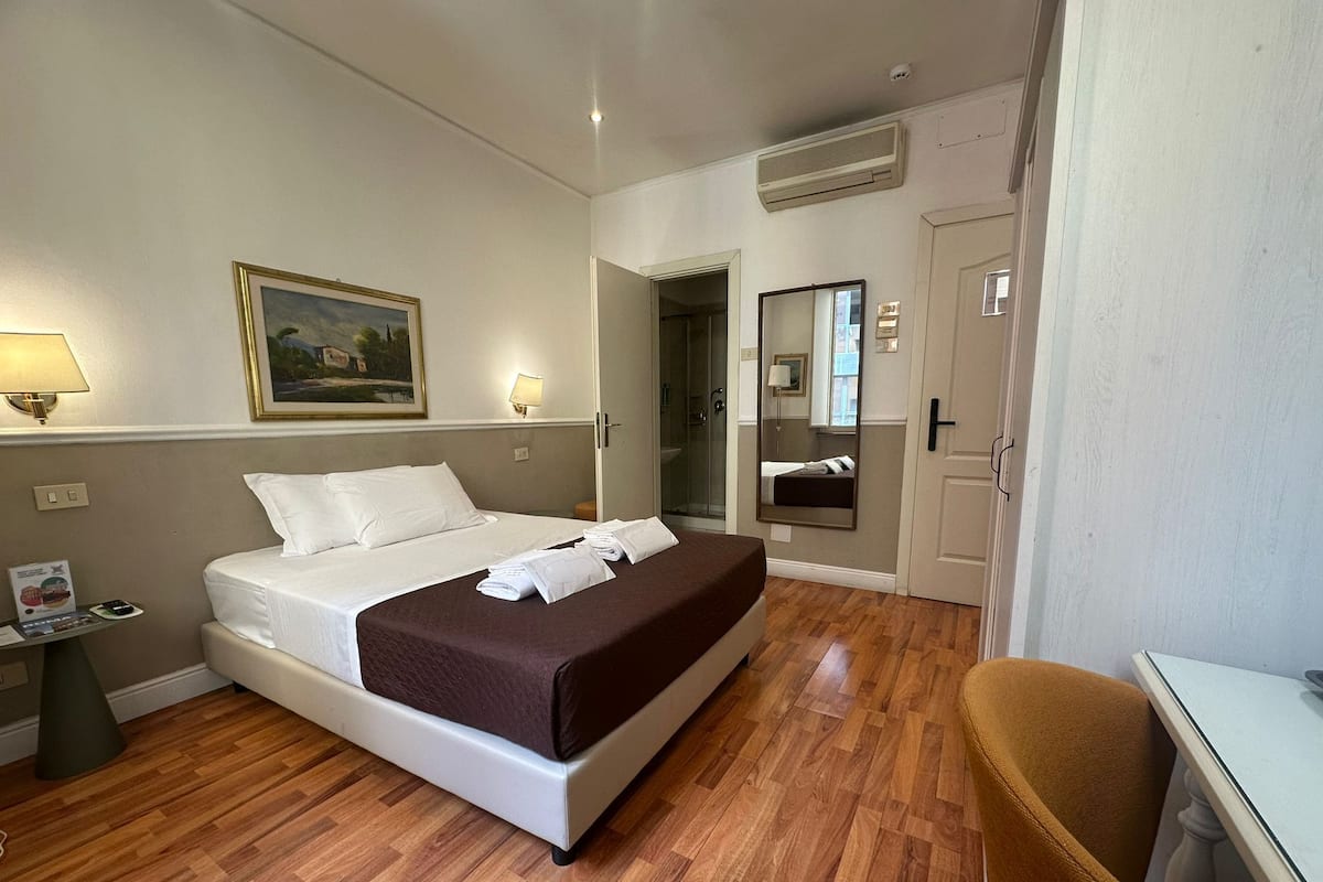 A hotel room with a bed, bedside table, lamp, painting, and a mirror.