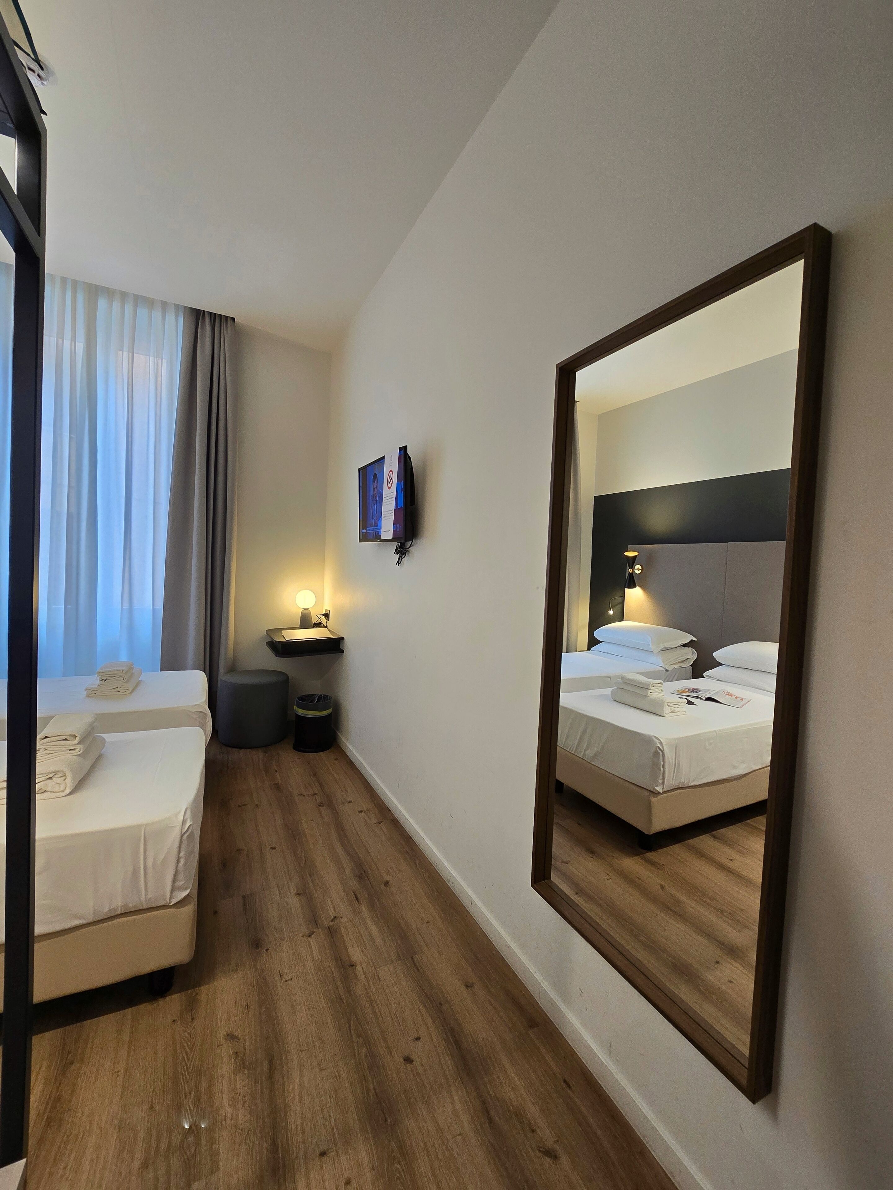 A hotel room with two beds, a TV mounted on the wall, a bedside table with a lamp, and a large mirror reflecting the room.