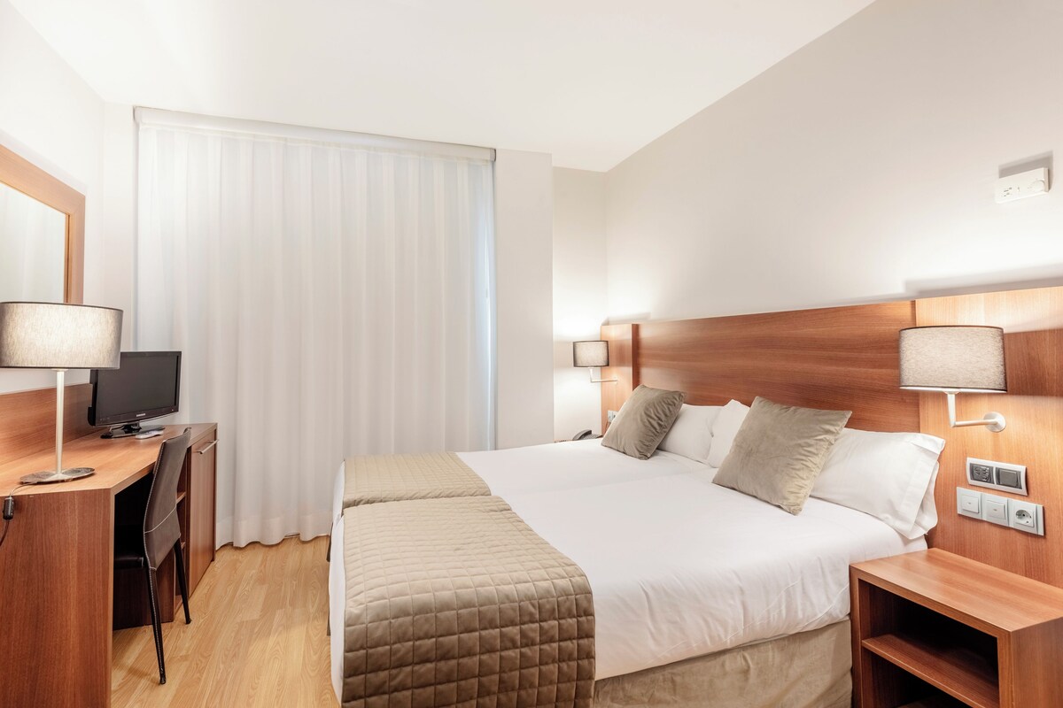 A hotel room with a bed, bedside tables, a desk with a chair, a television, and a window with curtains.