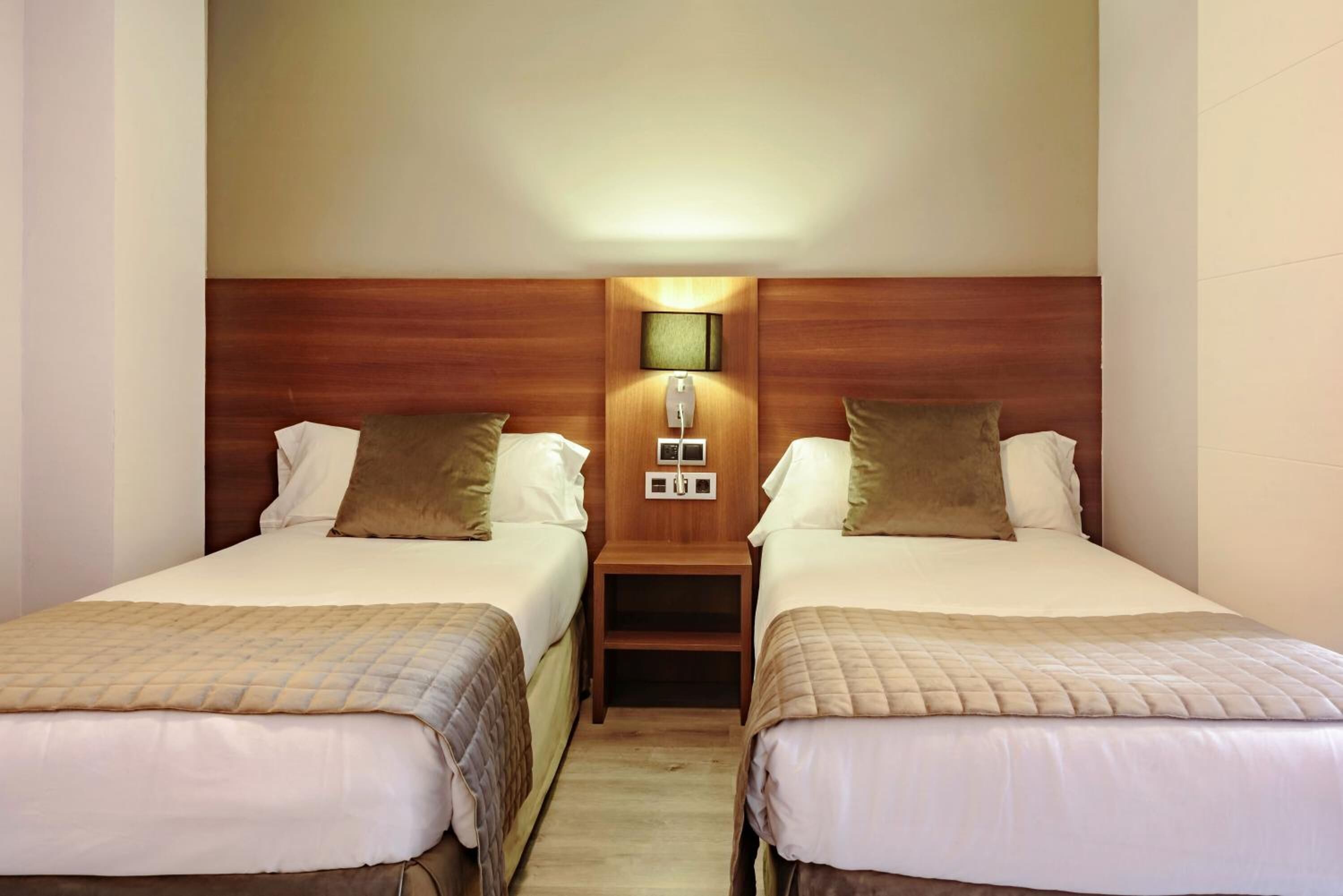 A hotel room with two beds, a nightstand with a lamp, and a wall-mounted light fixture.