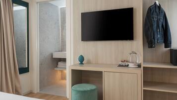 A hotel room with a TV mounted on the wall, a desk with a kettle, a small plant, and a hanging jacket.