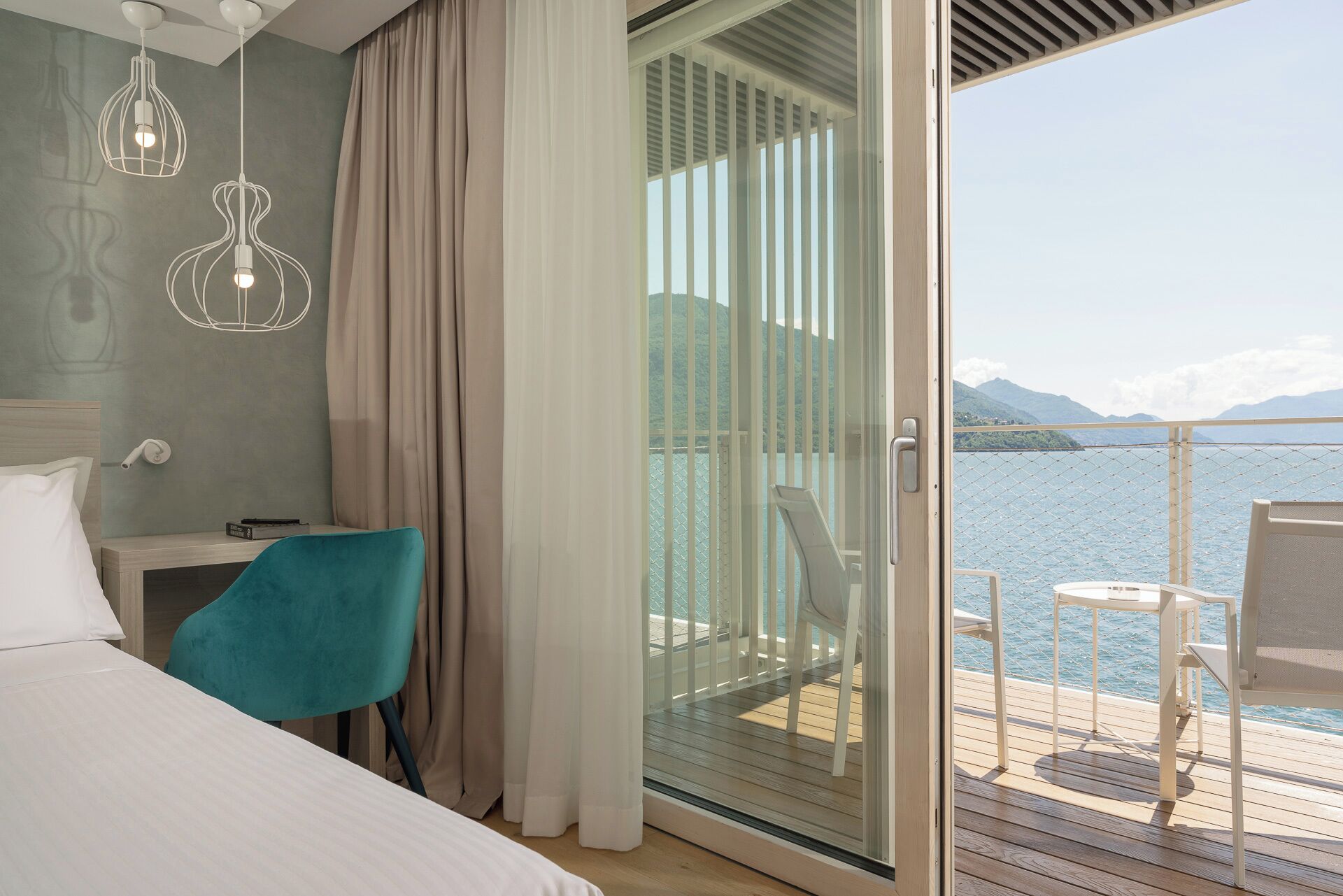 A bedroom with a bed, a teal chair, a nightstand, and a glass door leading to a balcony with a view of the sea and mountains.