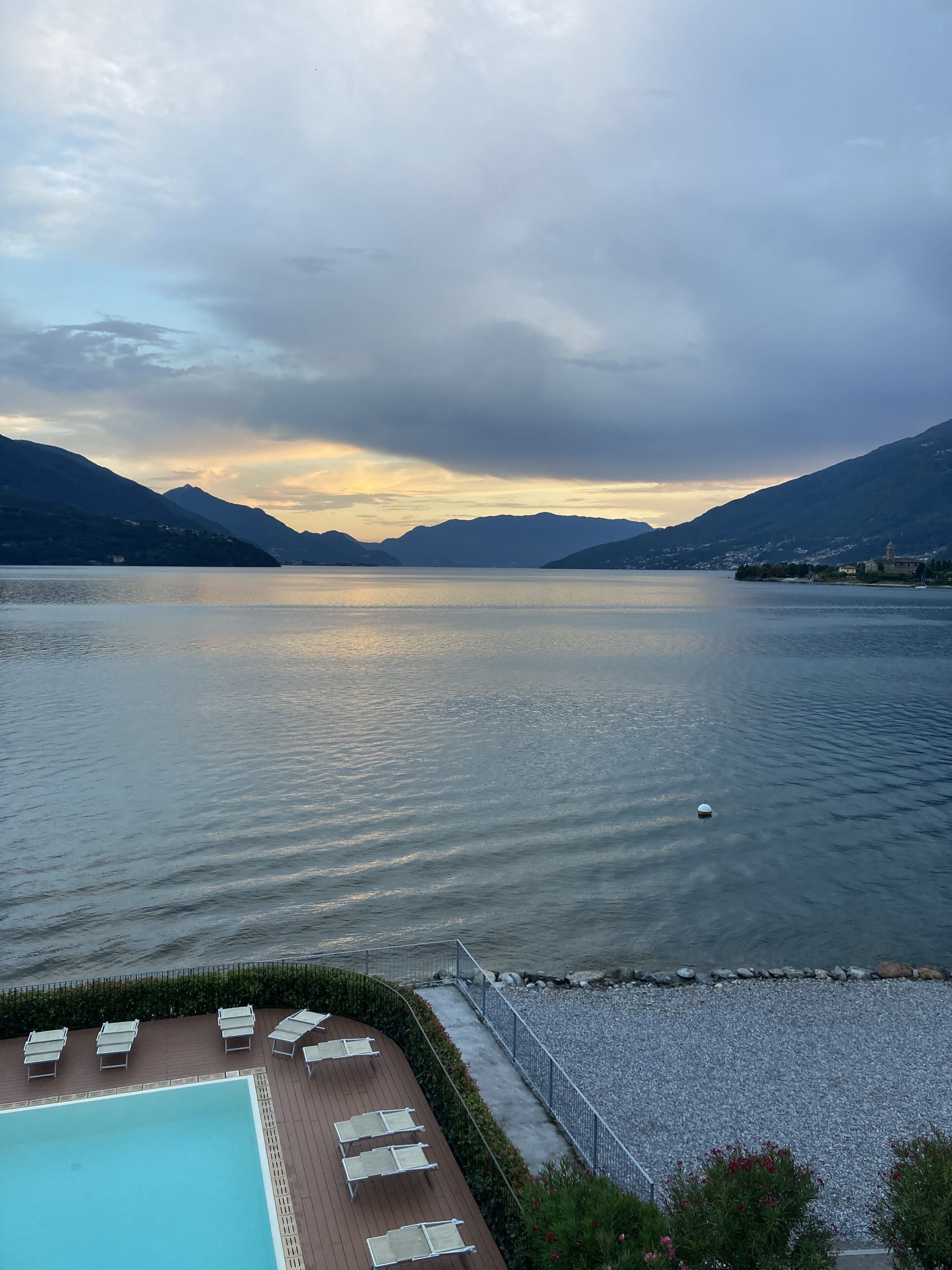 A swimming pool with lounge chairs, surrounded by a pebble beach and a view of a calm lake with mountains in the background.