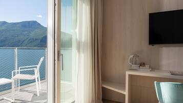 Un balcón con vistas al mar y a las montañas, una silla blanca, una mesa con hervidor y un televisor en la pared.