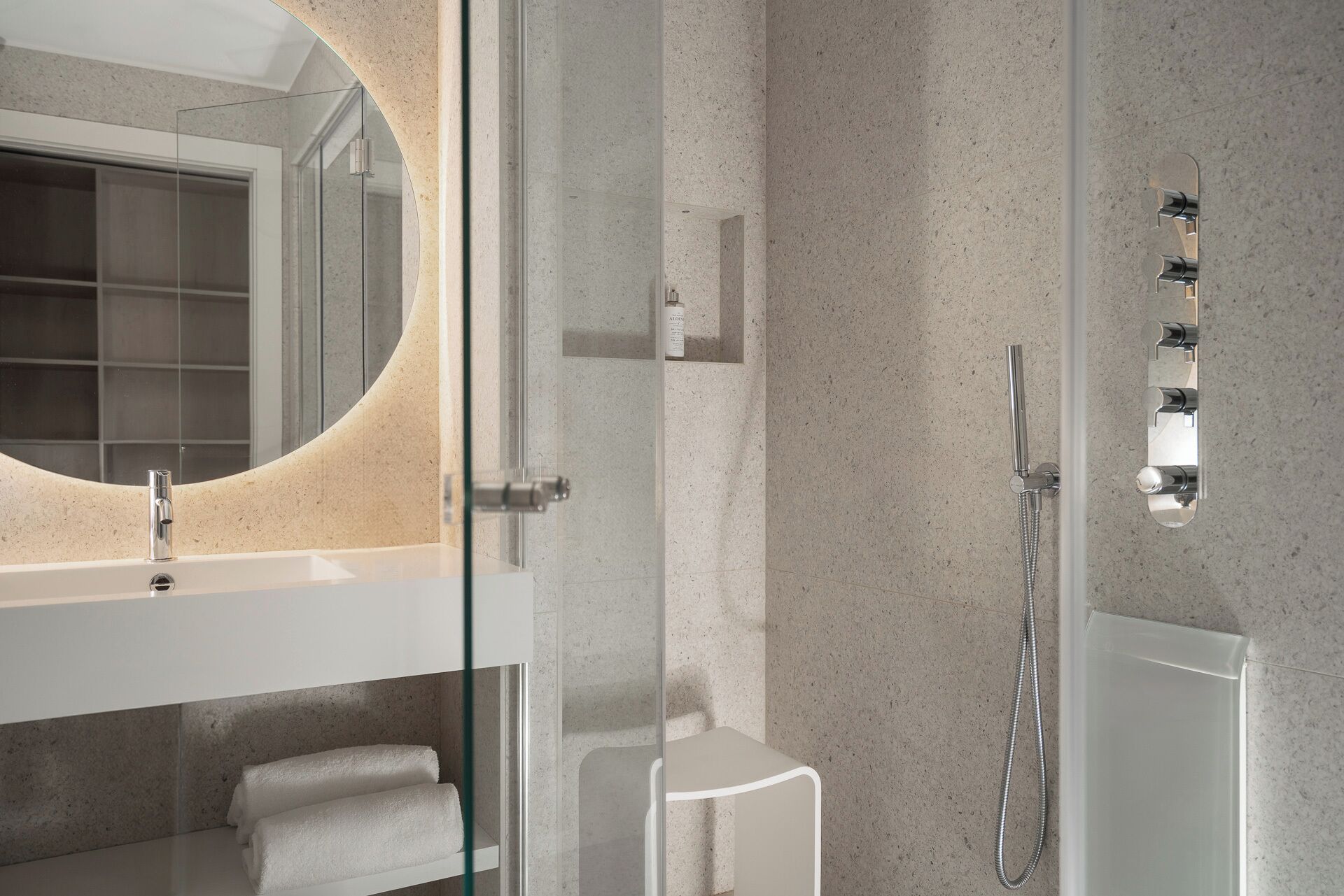 Un bany modern amb dutxa de vidre, un lavabo blanc i un mirall gran de marc circular.