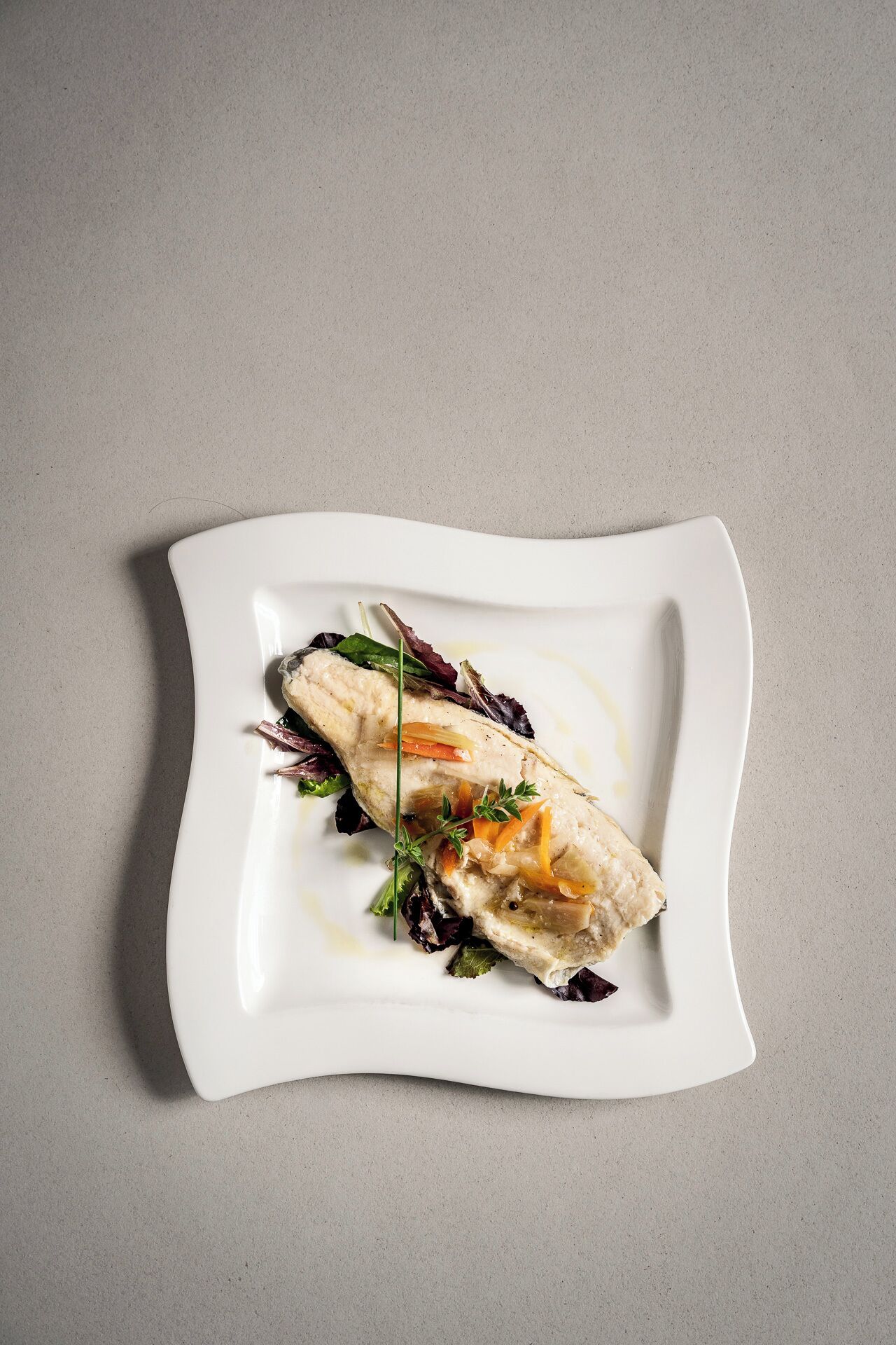 A white plate with a piece of fish and vegetables on a textured grey background.
