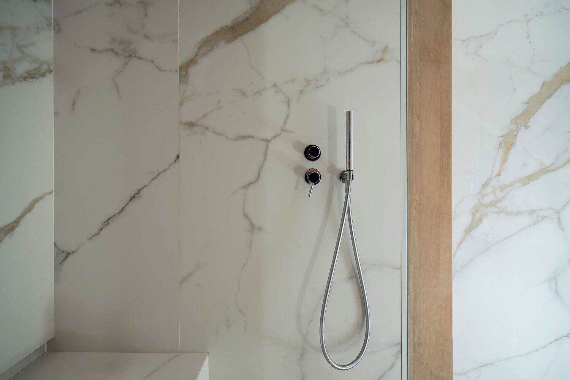 A bathroom with a marble wall, a shower with a handheld showerhead, and a wooden frame.