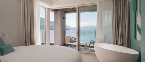 A bedroom with a large bed, a bathtub, and a balcony with a view of the water and mountains.