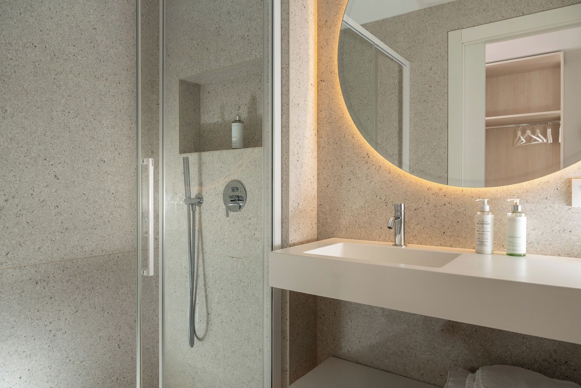 Un baño moderno con ducha envuelta en vidrio, un lavamanos blanco y un espejo con luz circular.