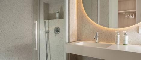 Un baño moderno con ducha envuelta en vidrio, un lavamanos blanco y un espejo con luz circular.