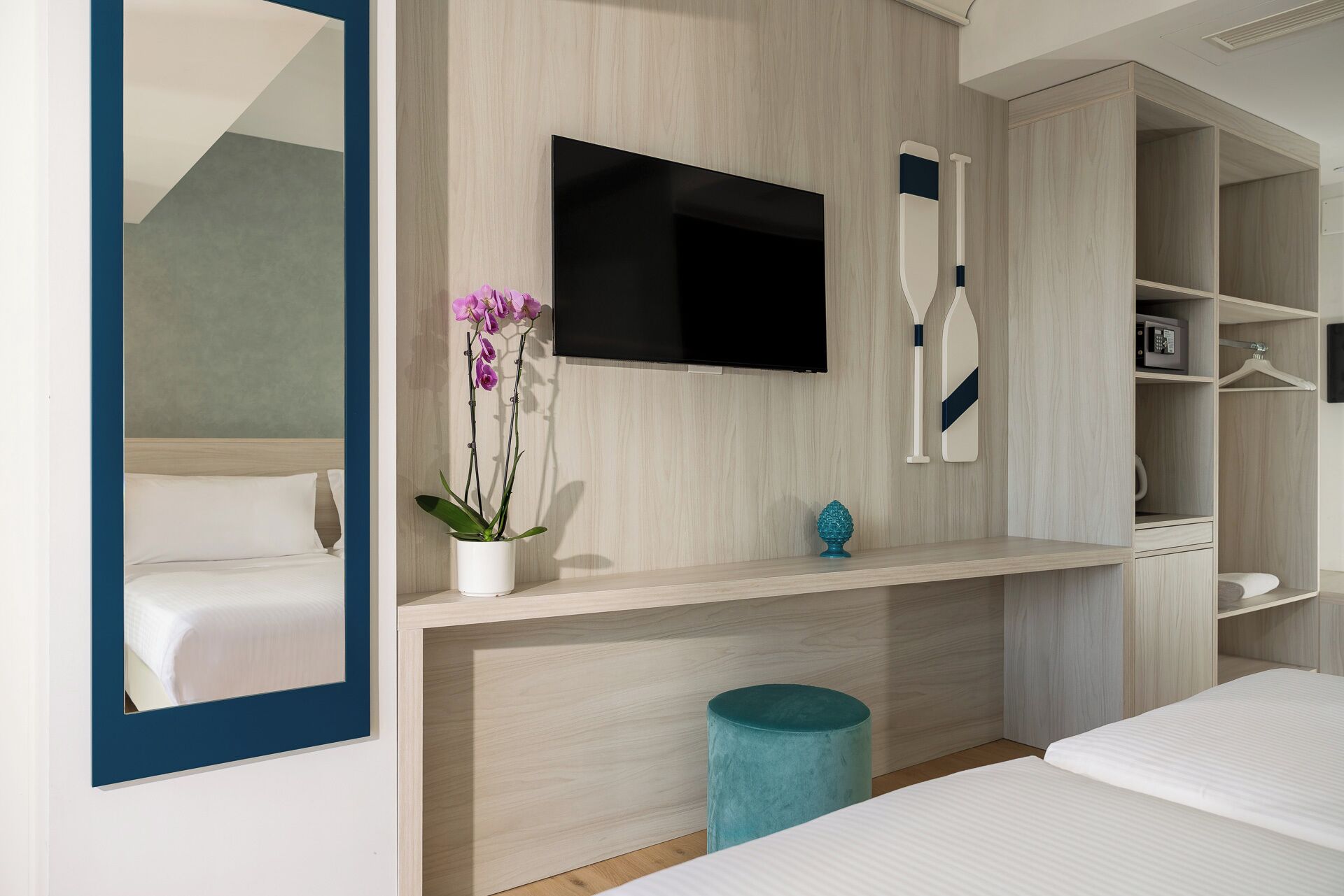 Una habitación de hotel moderna con una cama, un televisor montado en la pared, una planta pequeña y un reposapiés azul.