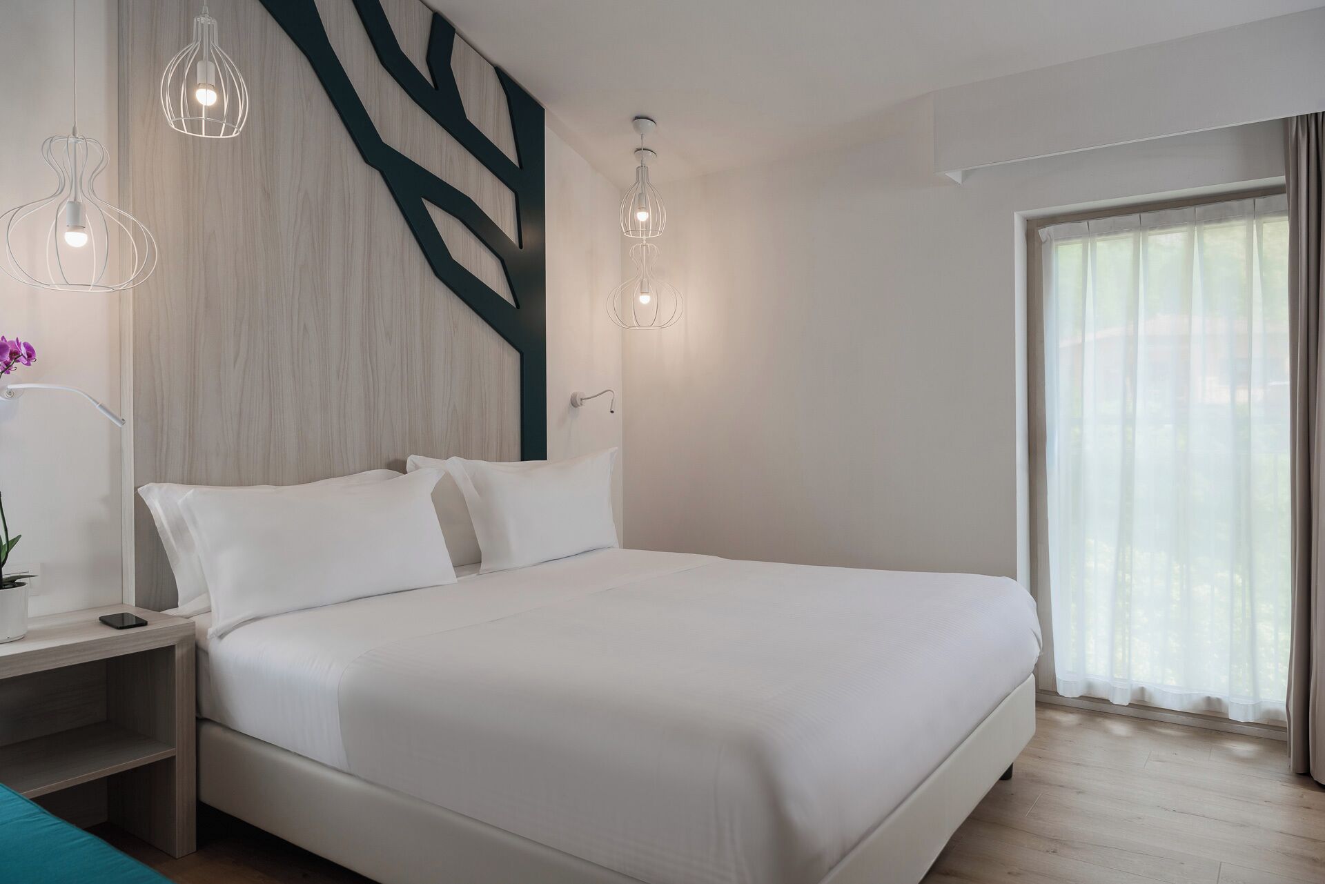 Une chambre d’hôtel moderne avec un grand lit, une table de chevet, un élément décoratif mural et une fenêtre avec des rideaux.