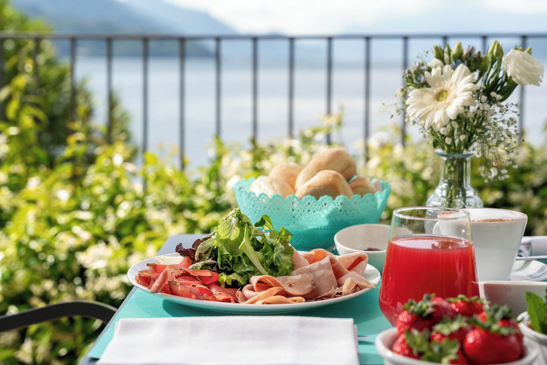 Un tavolo apparecchiato con un piatto di salumi, una ciotola di pane, un bicchiere di succo di frutta rossa e un vaso di fiori, con vista su un panorama suggestivo.