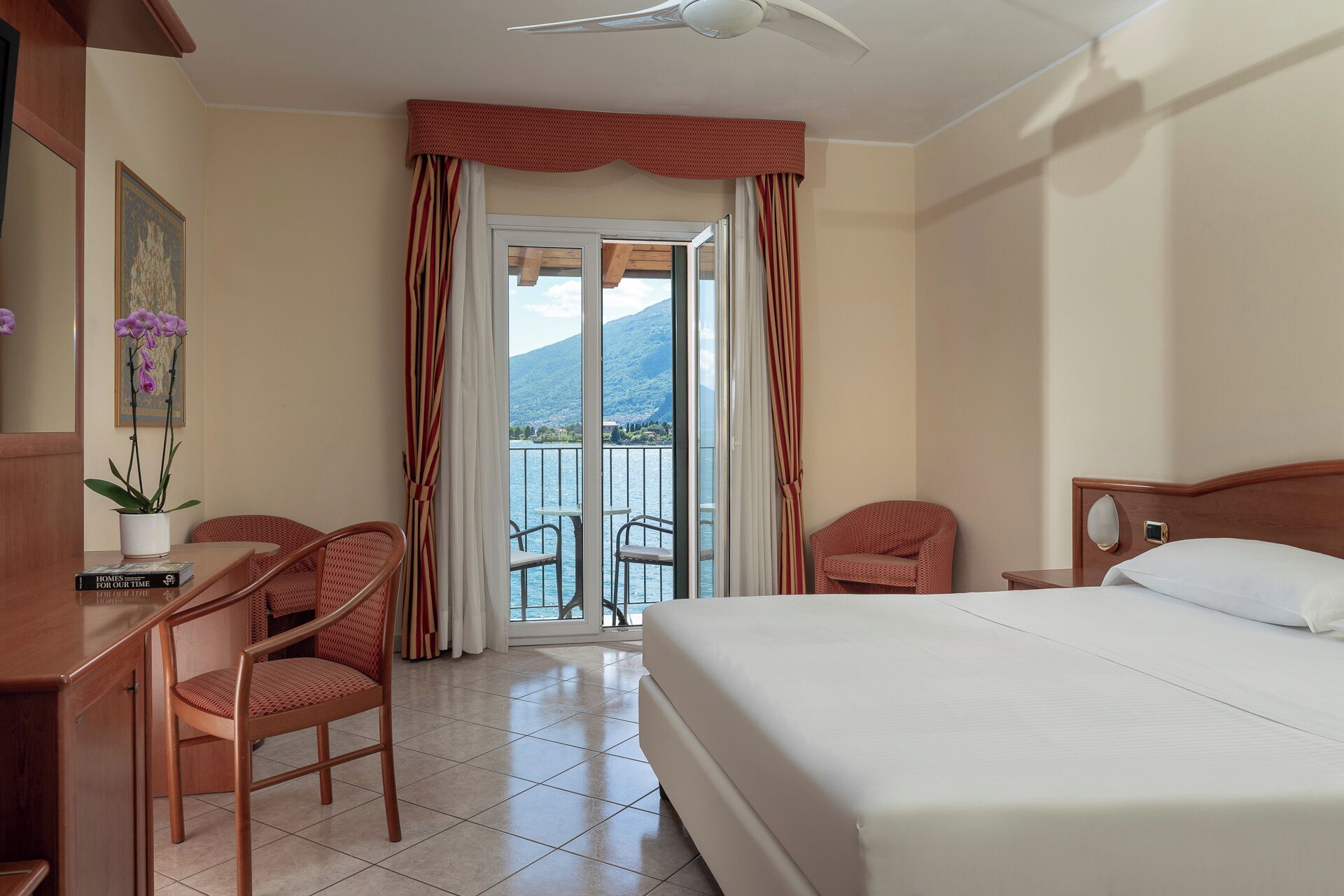 A hotel room with a bed, two chairs, a desk, a potted plant, and a view of a lake and mountains through a sliding door.