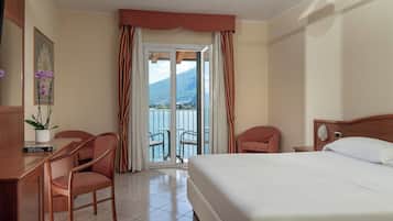 Habitación de hotel con cama, dos sillas, escritorio, planta en maceta y vista a un lago y montañas a través de una puerta corredera.