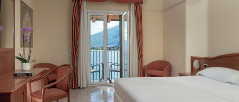 A hotel room with a bed, two chairs, a desk, a potted plant, and a view of a lake and mountains through a sliding door.