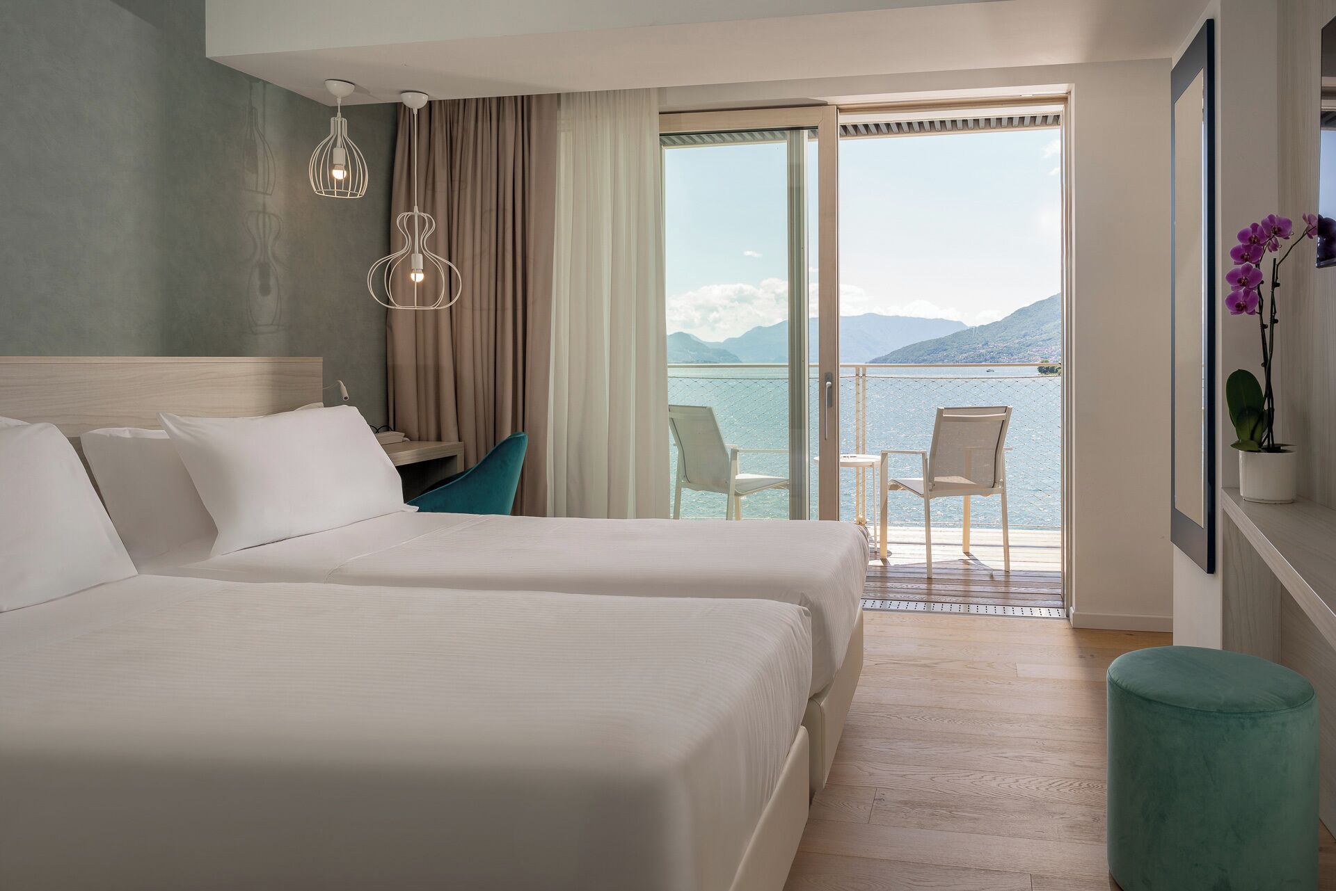 A hotel room with two beds, a desk, a chair, and a balcony with a view of the sea and mountains.