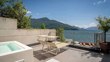 A terrace with a hot tub, lounge chairs, and a view of a lake with mountains in the background.