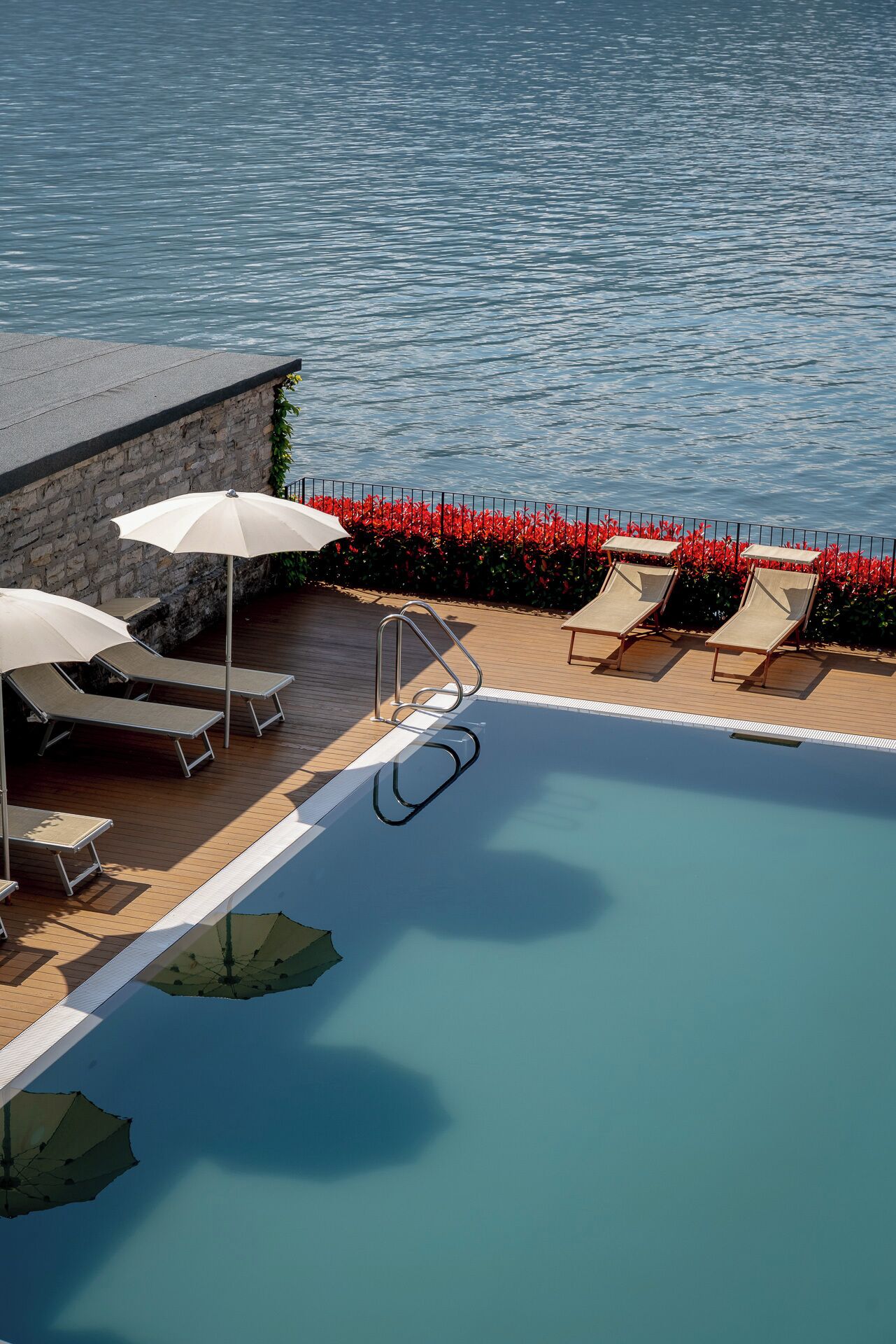 Ein Swimmingpool mit Liegestühlen und Sonnenschirmen, umgeben von einer Steinmauer und roten Blumen.
