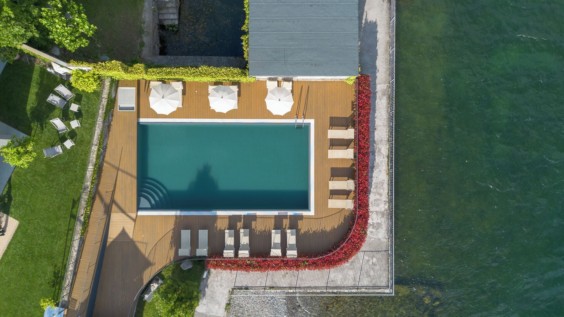 An aerial view of a rectangular swimming pool with a wooden deck, surrounded by lounge chairs and umbrellas, with a water body on one side.