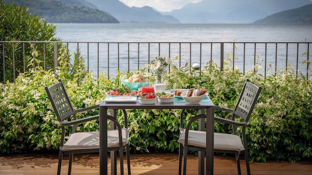 Een tafel gedekt met eten en drinken op een balkon met uitzicht op een meer, omgeven door groen en bergen.