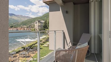 A balcony with a wicker chair and a table, overlooking a lake and a mountainous town.