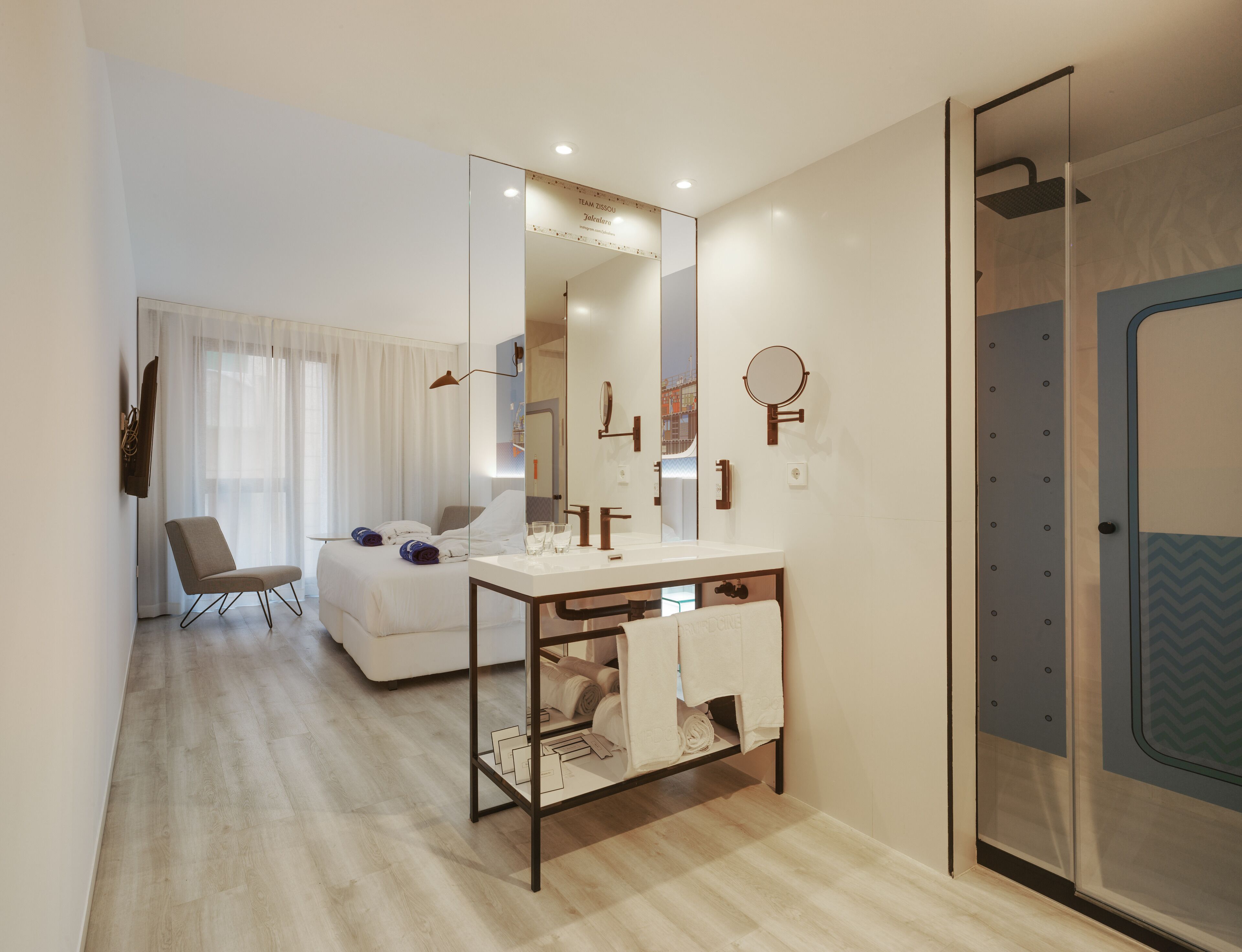A modern hotel room with a large bed, a bedside table, a mirror, a shower area, and a seating area with a chair and a TV.