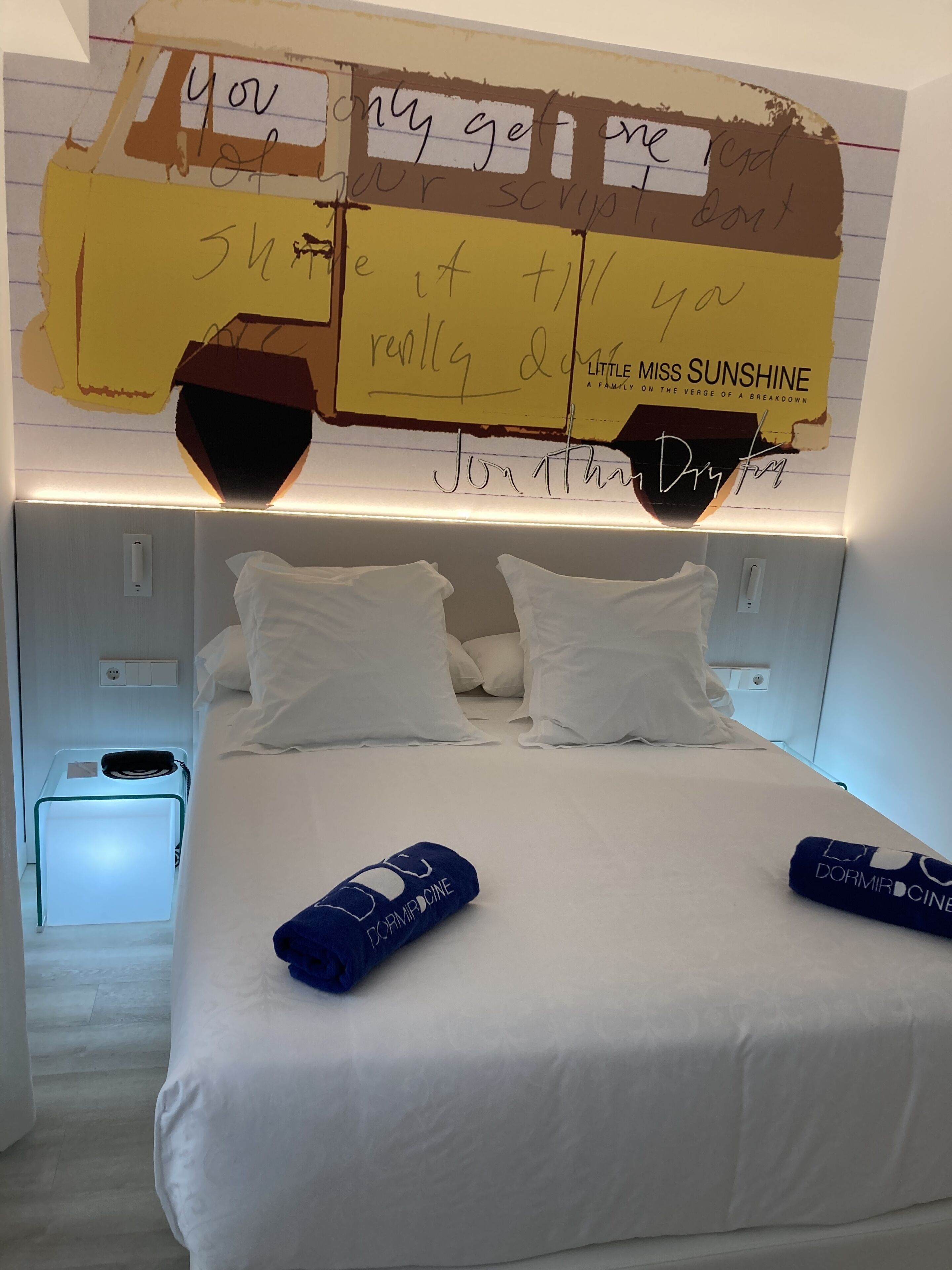 A hotel room with a neatly made bed, a bedside table, and a wall with a handwritten message.