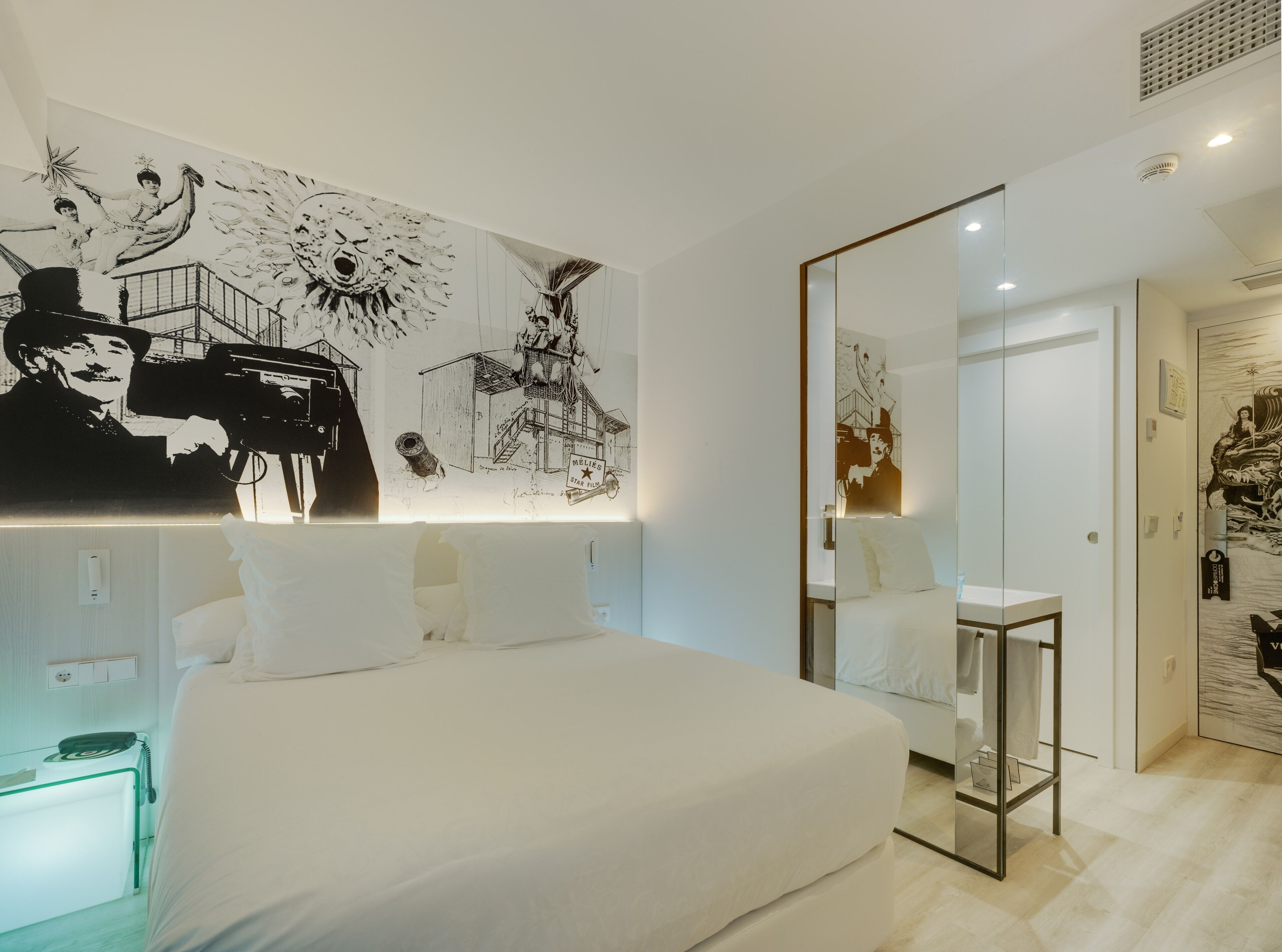 A modern hotel room with a large bed, a bedside table, and a wall with black and white artwork.
