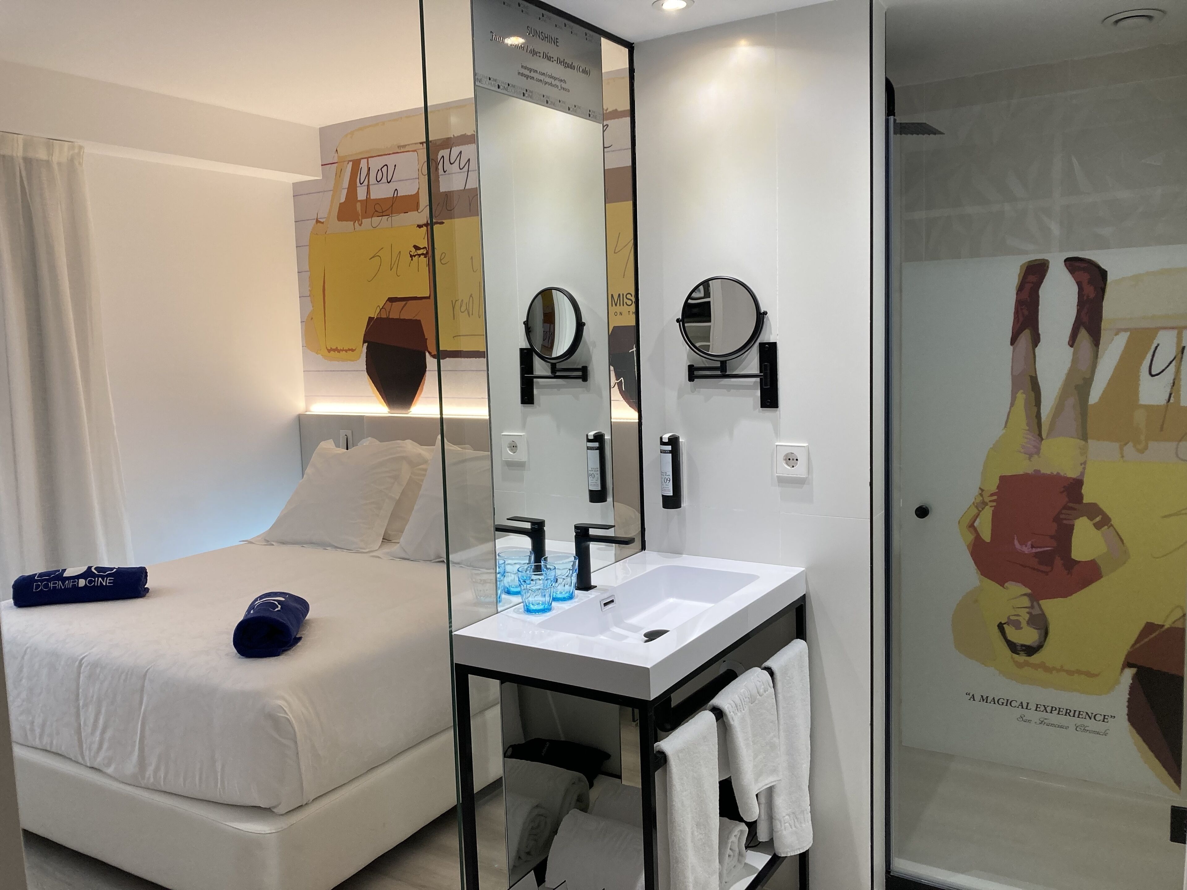 Une chambre d’hôtel moderne avec un lit, une table de chevet dotée d’un miroir, un lavabo blanc et un espace douche.