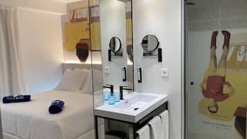A modern hotel room with a bed, a bedside table with a mirror, a white sink, and a shower area.