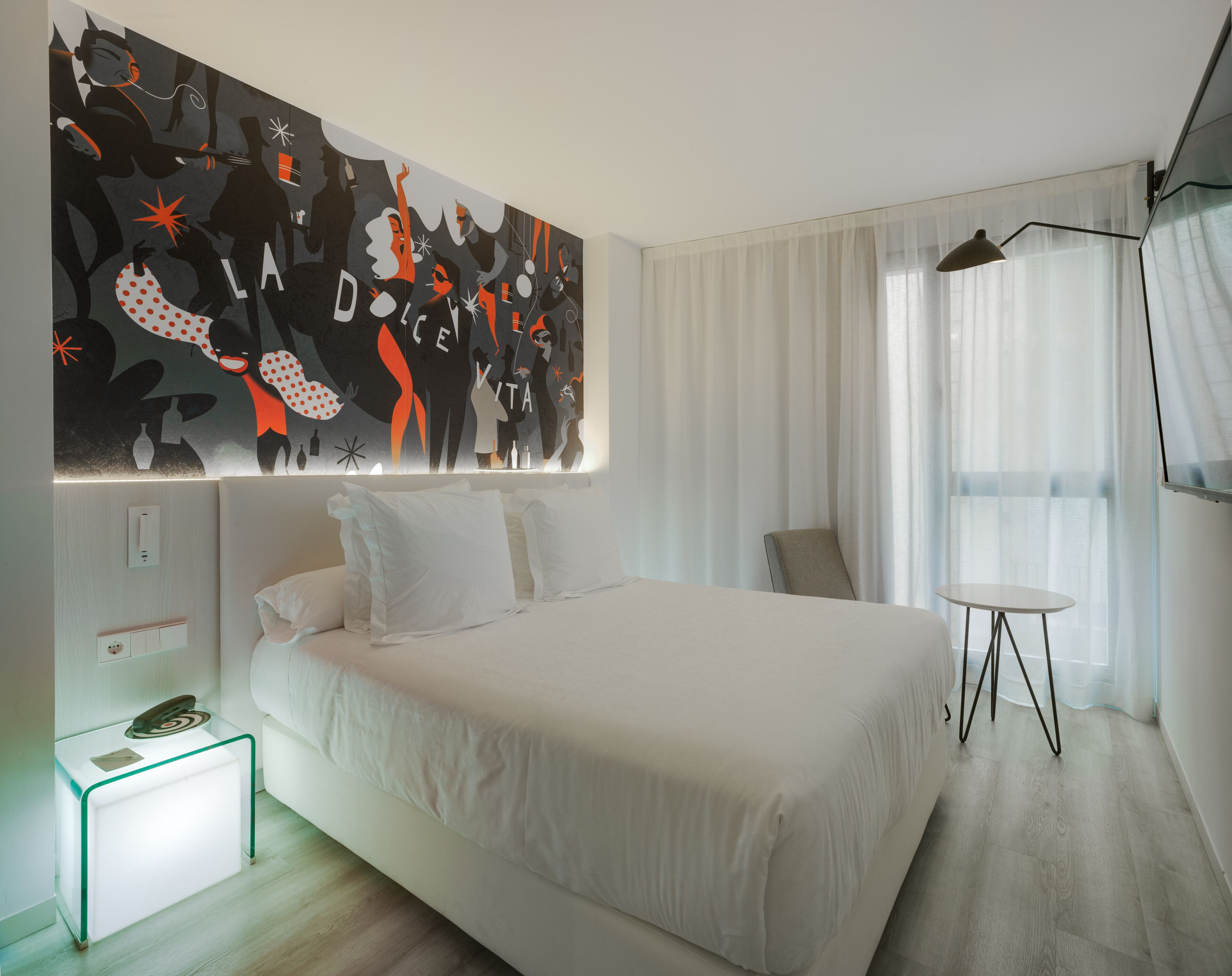 A modern hotel room with a large abstract painting on the wall, a bed with white linens, a bedside table with a lamp, and a view of the city through sheer curtains.