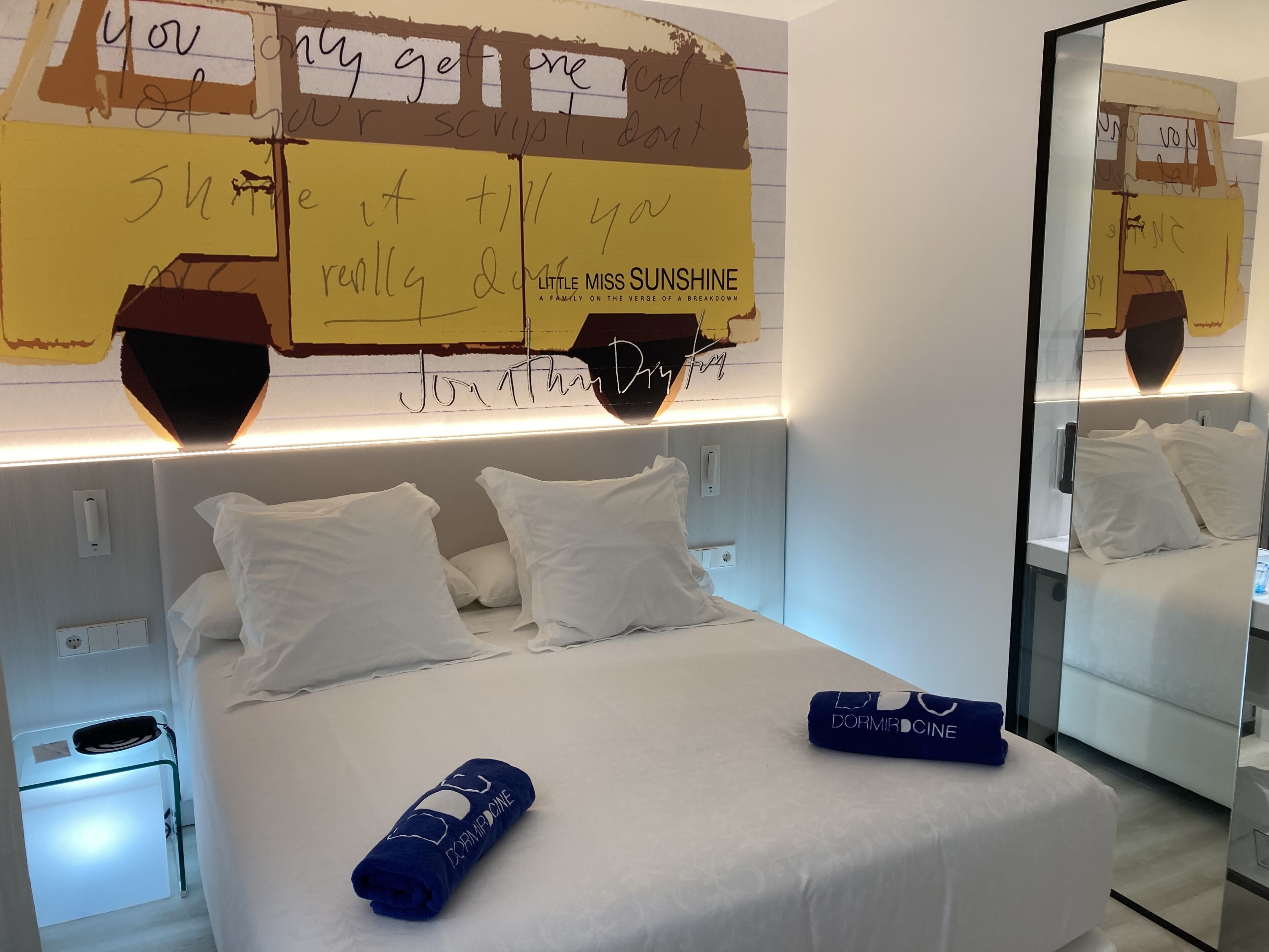 A hotel room with a neatly made bed, a bedside table with a lamp, and a wall with handwritten notes.