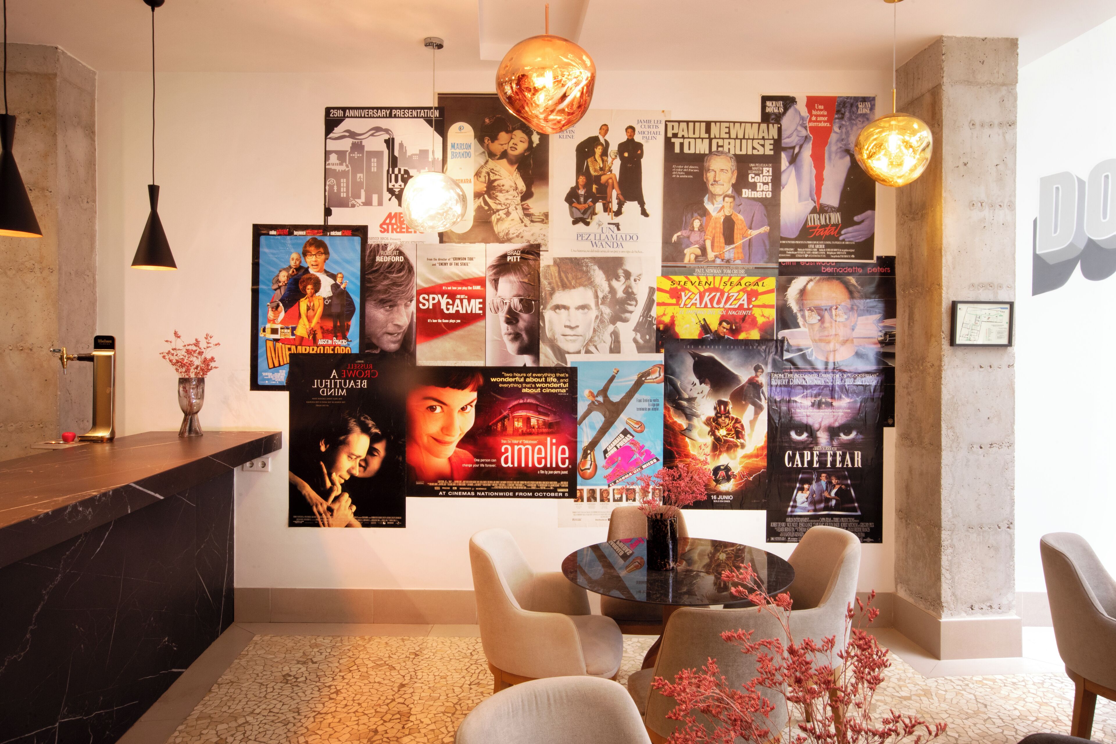 A lobby with a wall decorated with movie posters, a round table with chairs, and a reception desk with a vase of flowers.