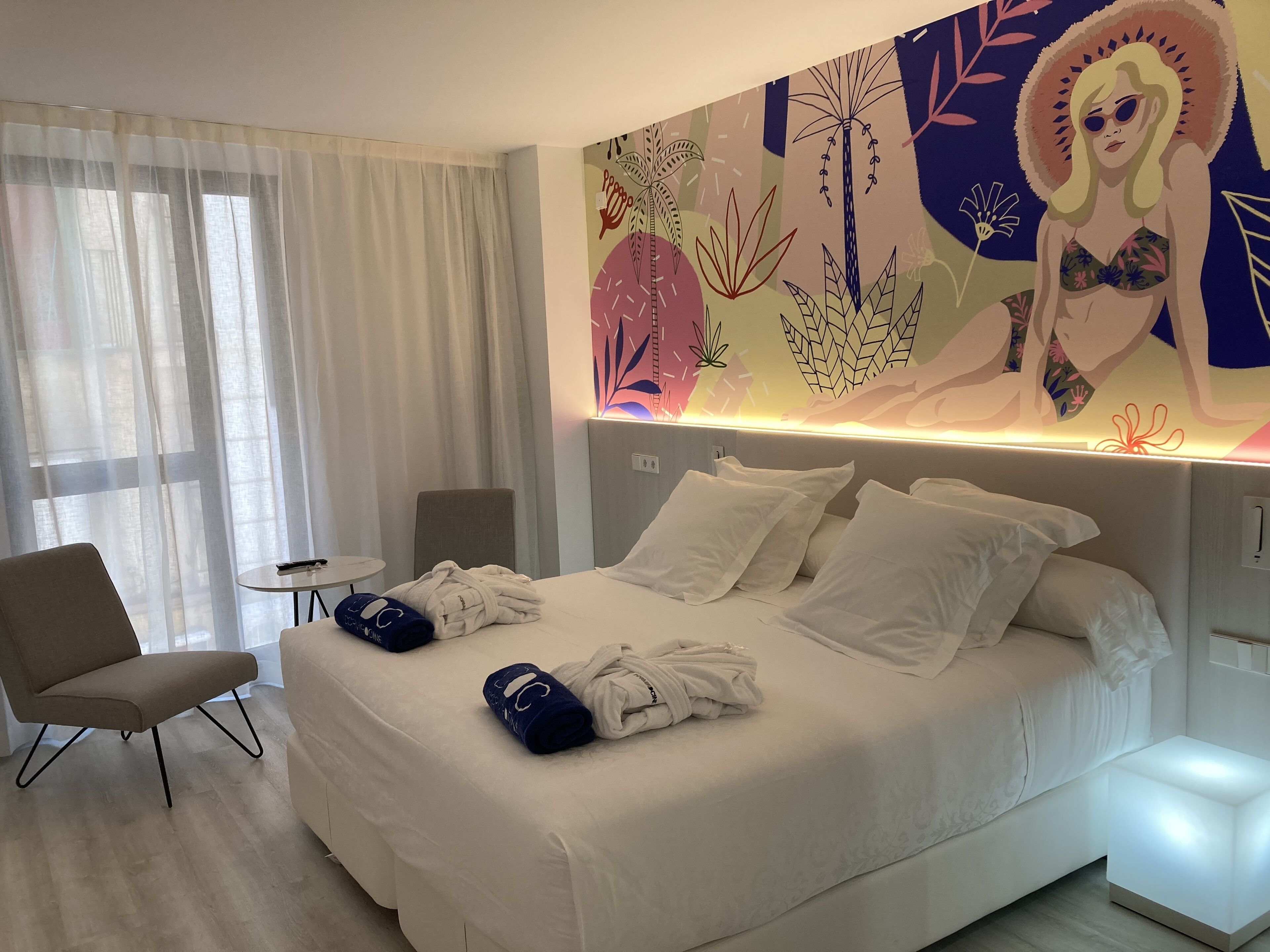 A hotel room with a large bed, a chair, a small table, and a wall mural.