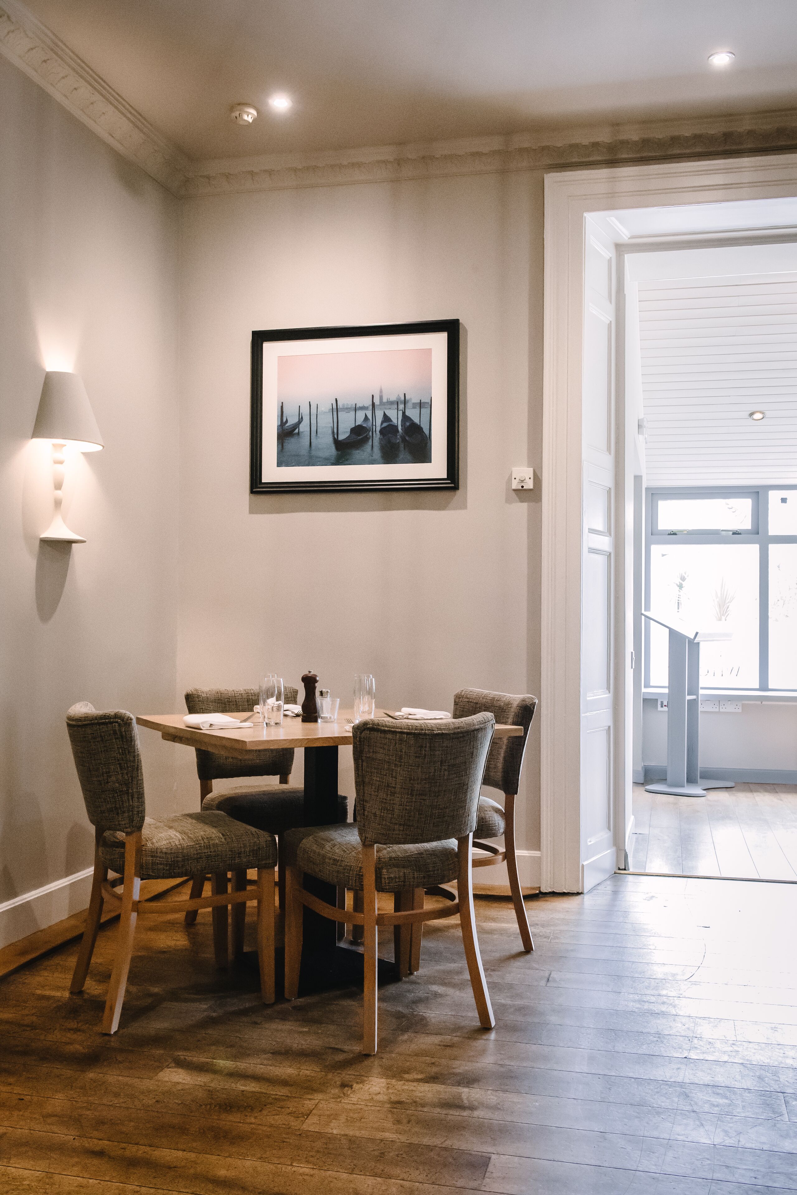 A dining area with a round table, four chairs, a framed picture on the wall, and a lamp on the wall.