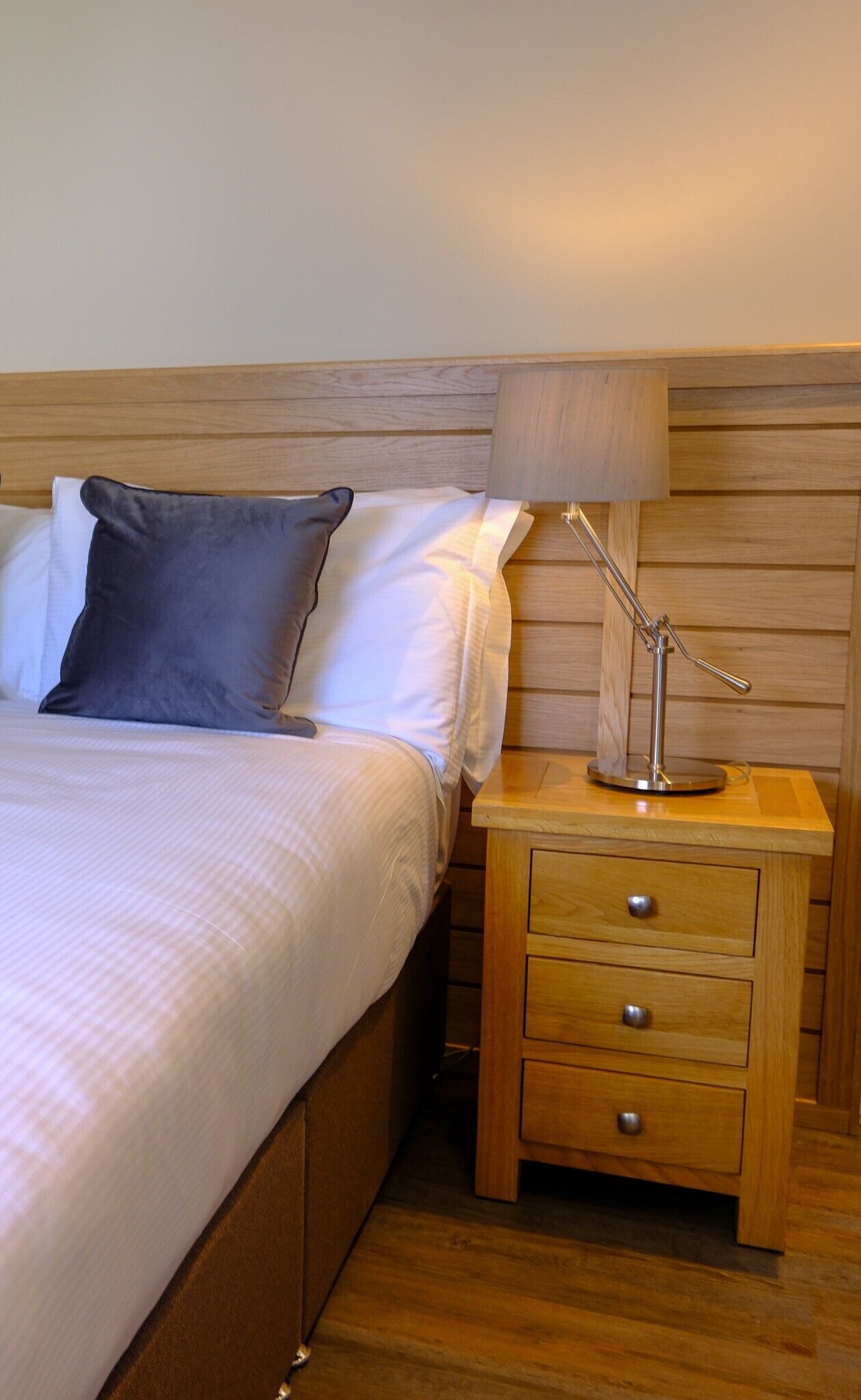 A neatly made bed with white and gray linens, a wooden headboard, and a bedside table with a lamp and drawers.