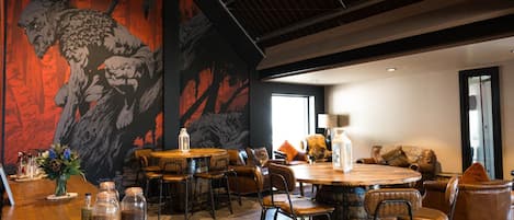 A modern dining area with a large mural, wooden tables, leather chairs, and a cozy seating area with cushions.