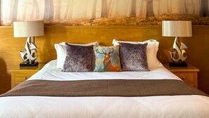 A hotel room with a large forest-themed mural on the wall, a neatly made bed with a brown throw blanket, and two nightstands with lamps and decorative items.