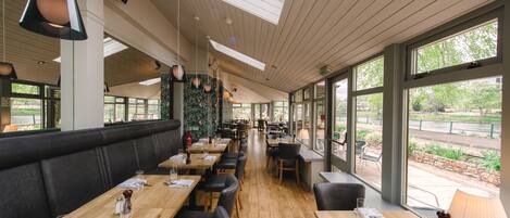 Ein Restaurant mit Holztischen, schwarzen Polsterbänken und großen Fenstern mit Blick ins Freie.