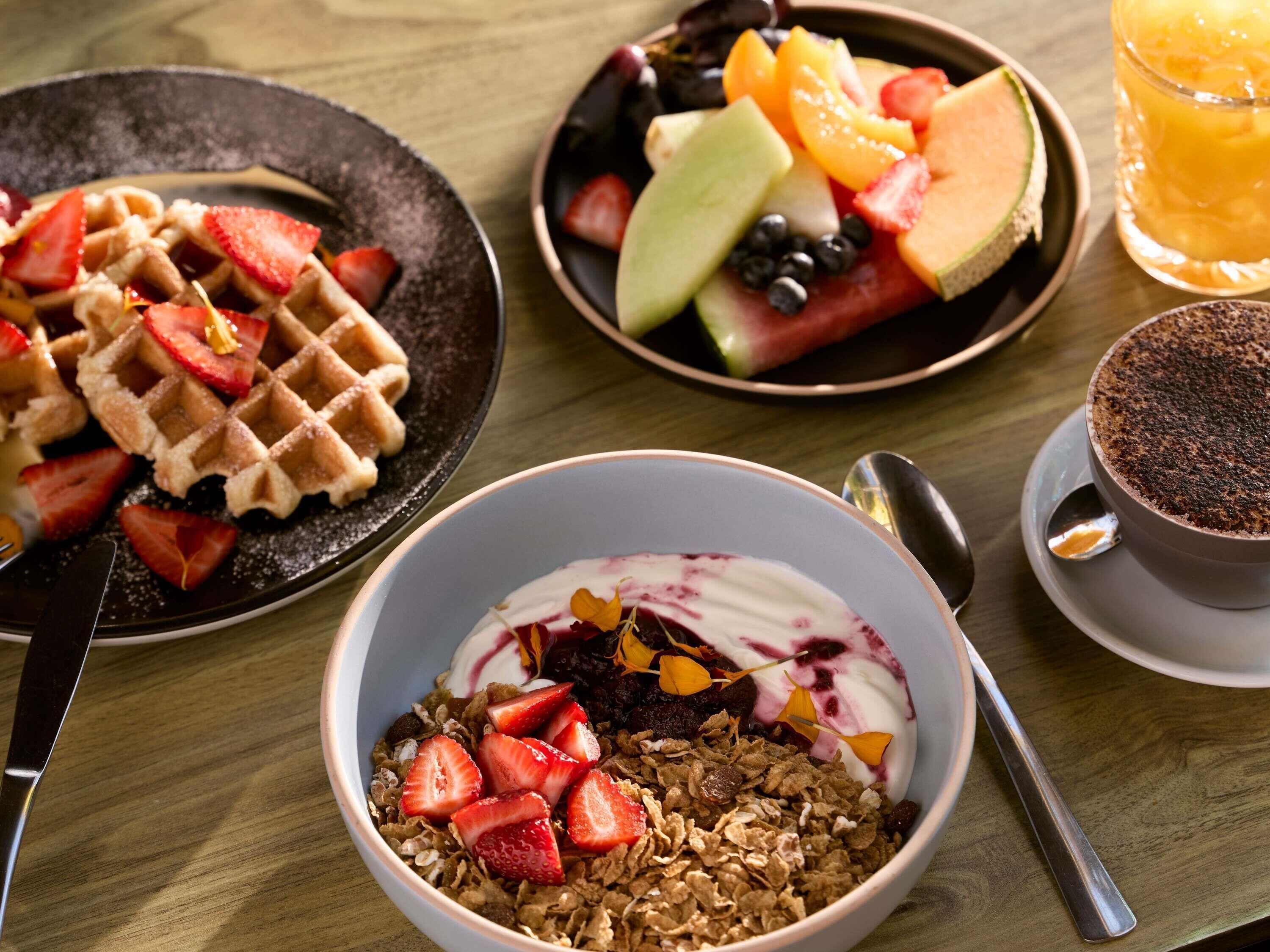 A breakfast setting with waffles, a bowl of yogurt with strawberries, a fruit plate, and a cup of coffee.