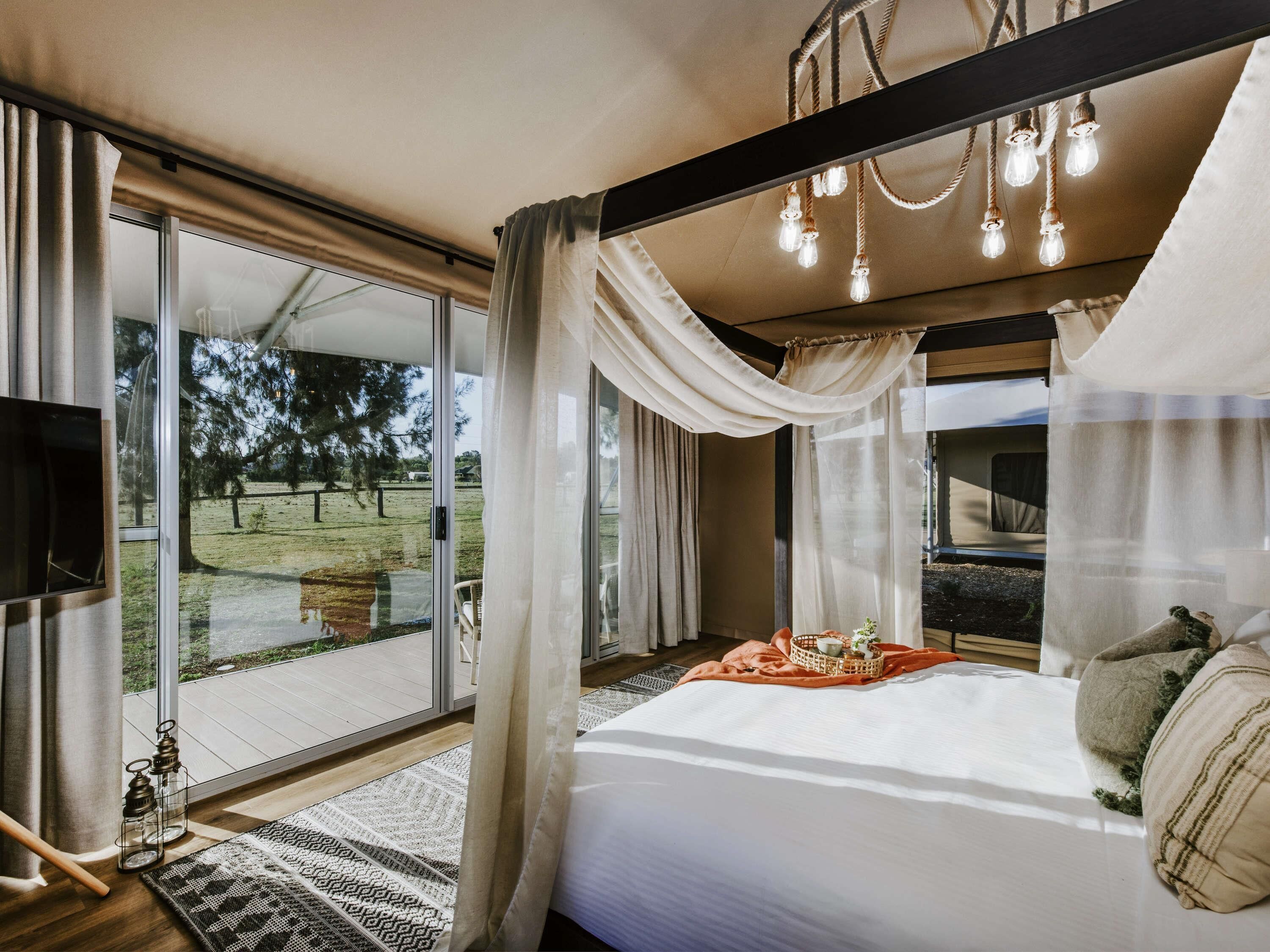 A bedroom with a large bed, a view of a grassy area, and a ceiling with hanging lights.