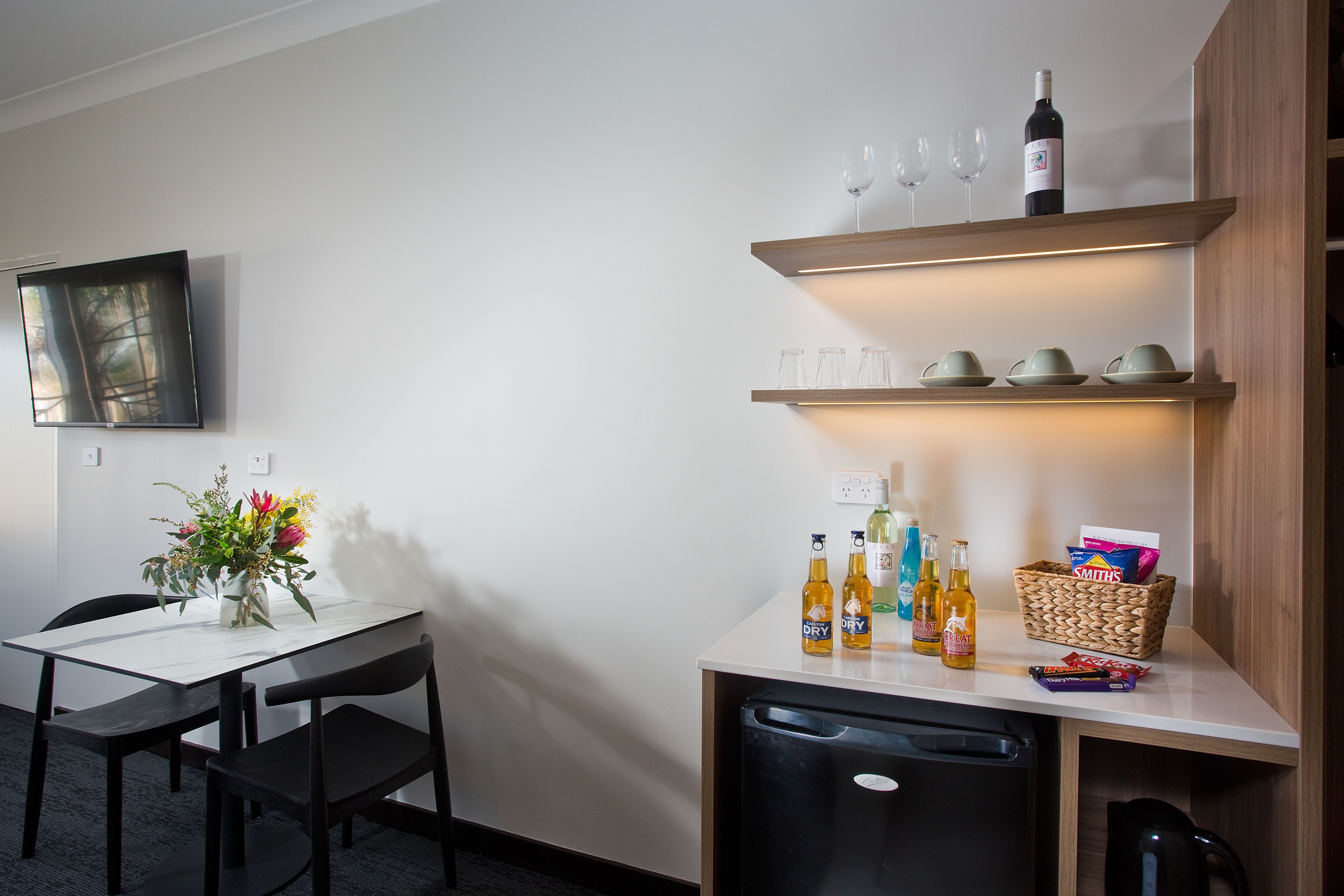 A modern kitchen with a white countertop, a dining table with chairs, a refrigerator, and a shelf with wine glasses and bottles.
