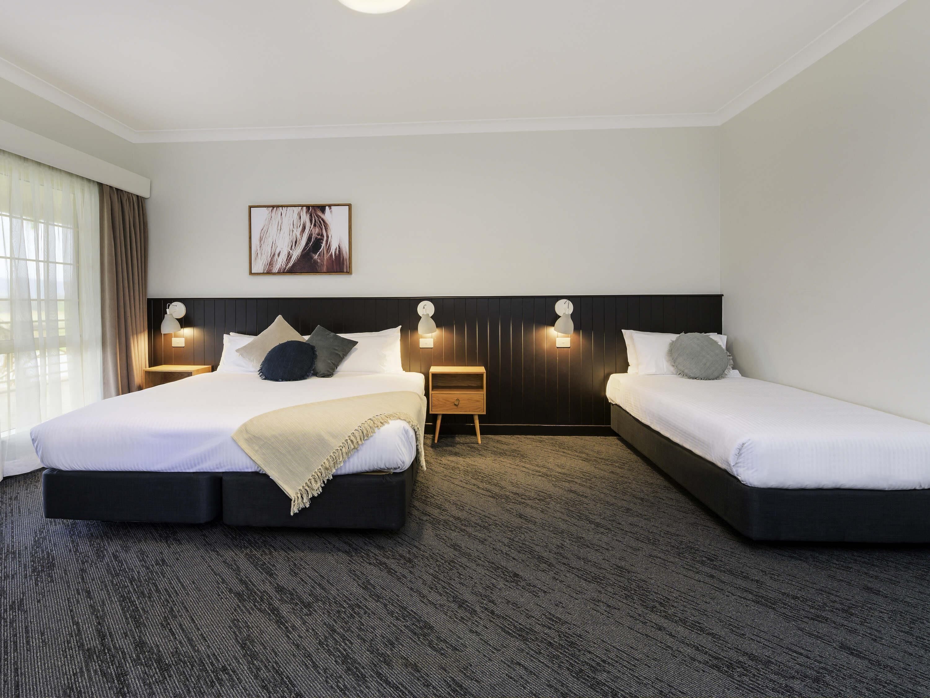 Une chambre d’hôtel avec deux lits, une grande fenêtre avec des rideaux, une horloge murale et un tableau au-dessus des lits.