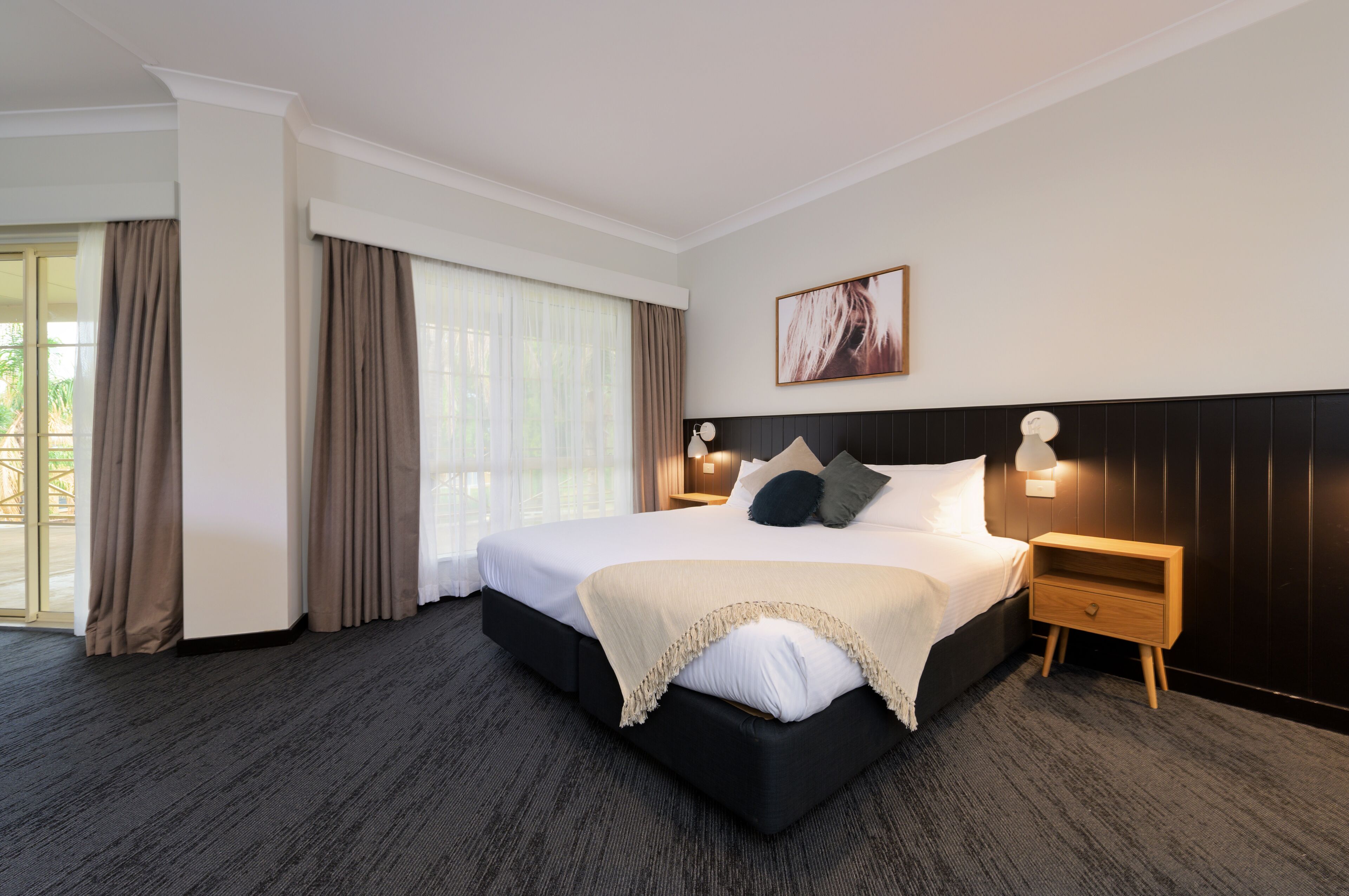 A hotel room with a large bed, a nightstand, a wall-mounted lamp, and a window with curtains.