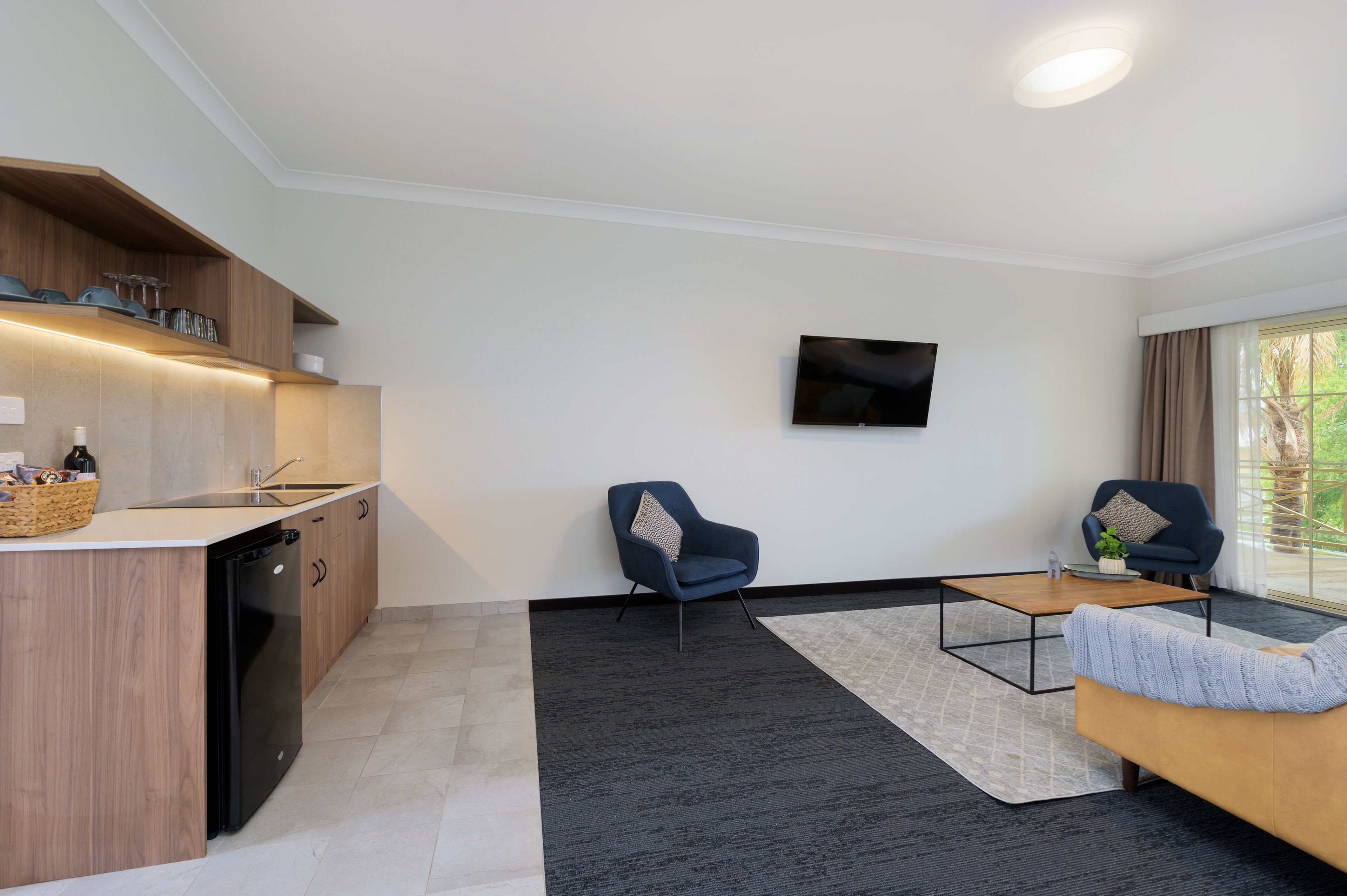 Un salon moderne avec un coin cuisine, un téléviseur à écran plat fixé au mur et une grande fenêtre avec des rideaux.