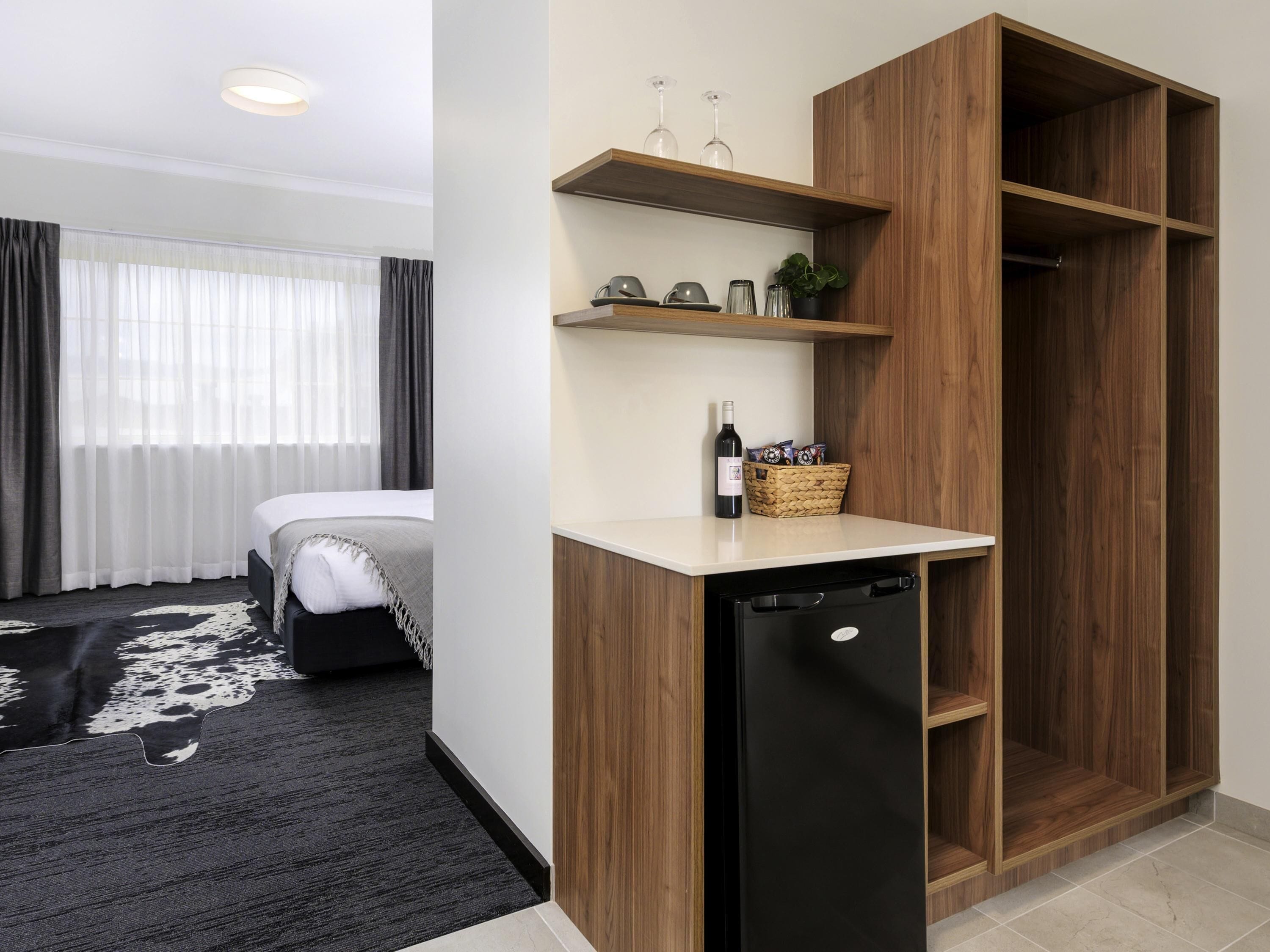 Una habitación de hotel compacta con cama, una pequeña cocina americana con refrigerador, un mueble de madera y una ventana con cortinas.