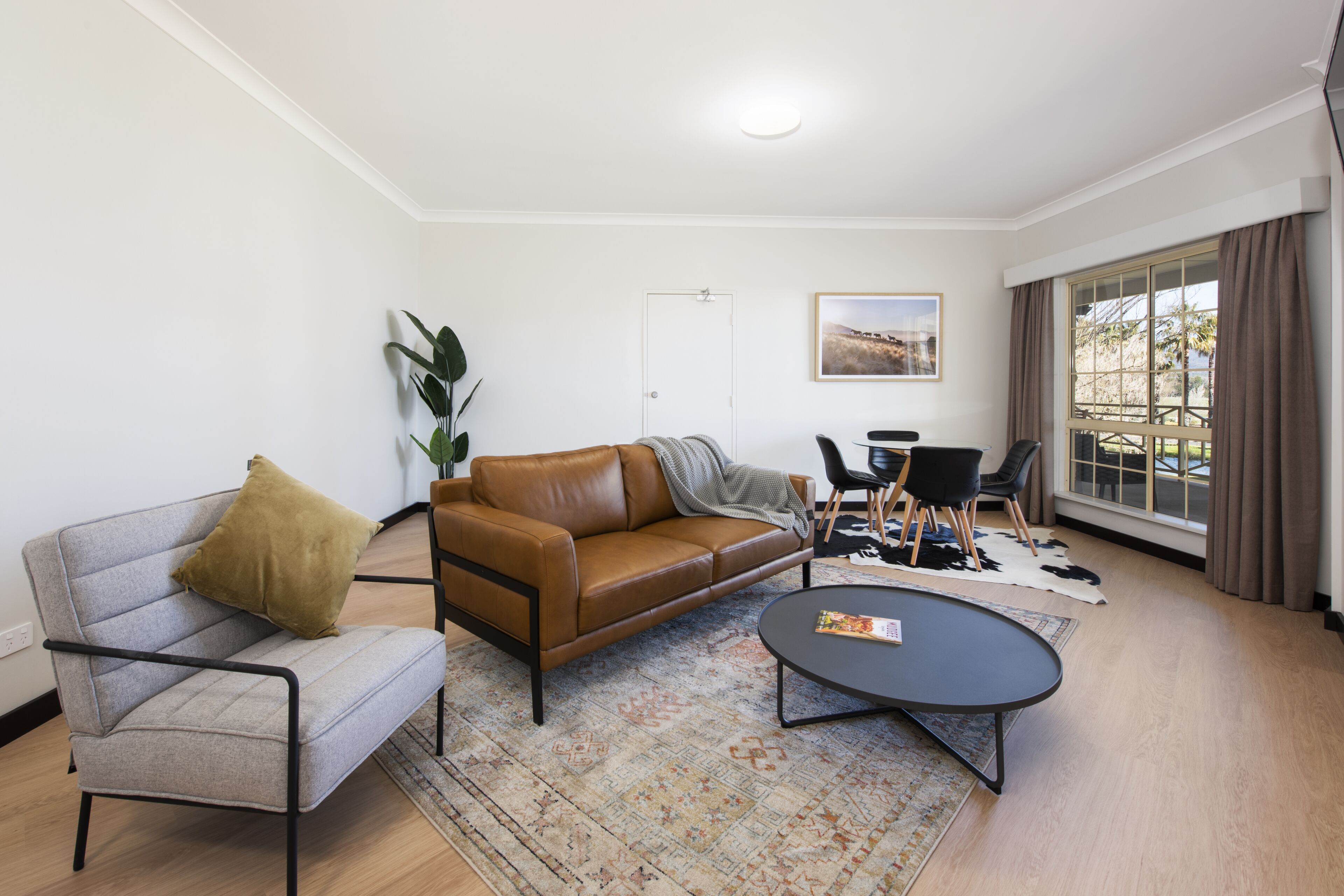 Una sala de estar moderna con un sofá de cuero marrón, un sillón gris, una mesa de centro redonda y un comedor con dos sillas.